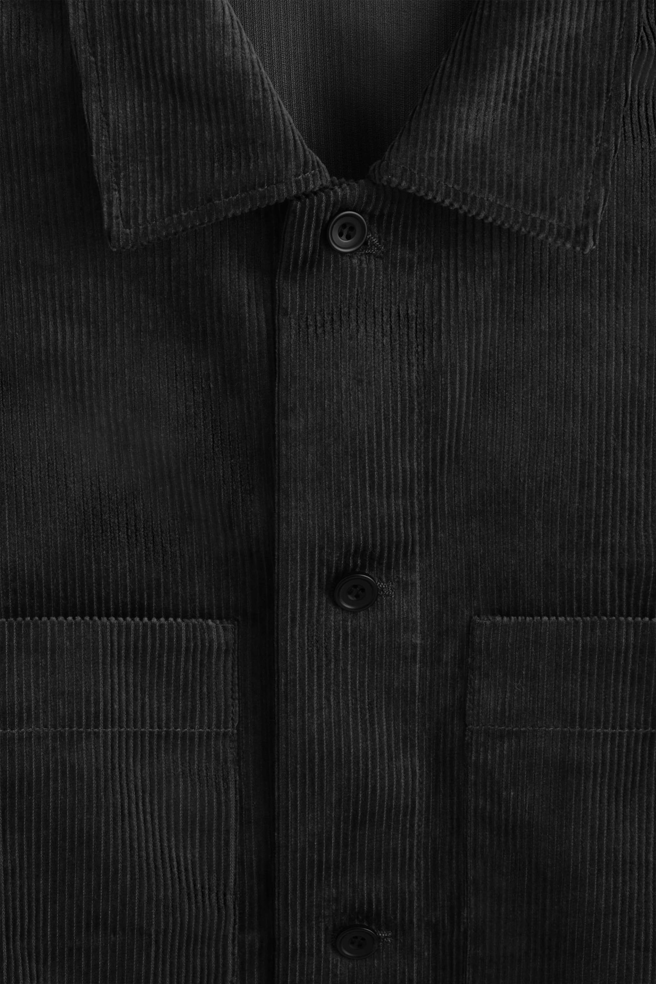 A black cotton corduroy overshirt is shown in a close-up front view, displaying its fine-ribbed texture, classic spread collar, and button-down front. Two front patch pockets sit on either side of the button placket, highlighting the garment’s utilitarian design. A black cotton corduroy overshirt is shown in a close-up front view, displaying its fine-ribbed texture, classic spread collar, and button-down front. Two front patch pockets sit on either side of the button placket, highlighting the garment’s utilitarian design.