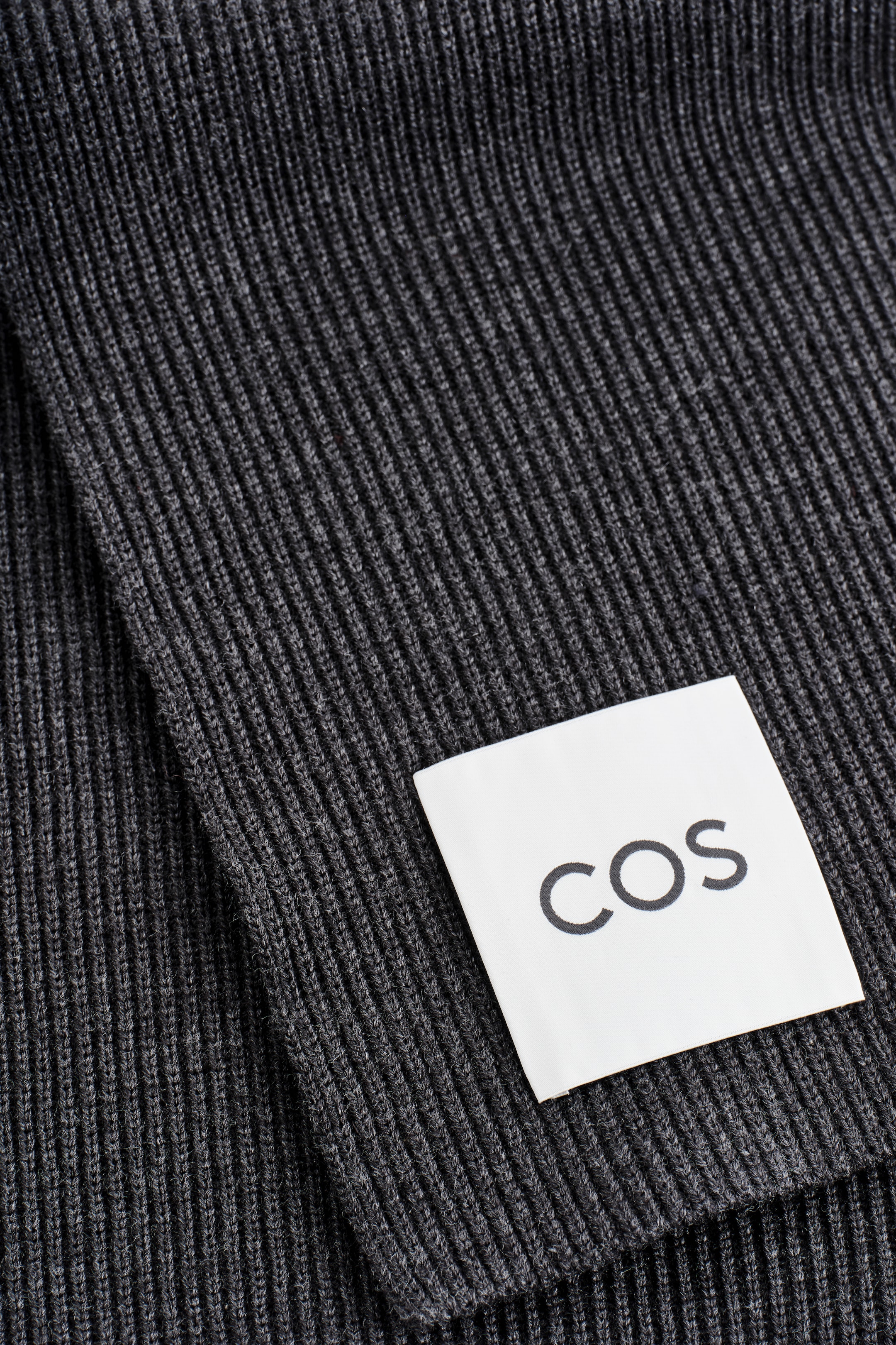 Ingrandisci l'immagine: Una sciarpa color carbone presenta una texture a maglia grossa a coste. Un'etichetta rettangolare bianca con "COS" in lettere nere è attaccata alla sciarpa.