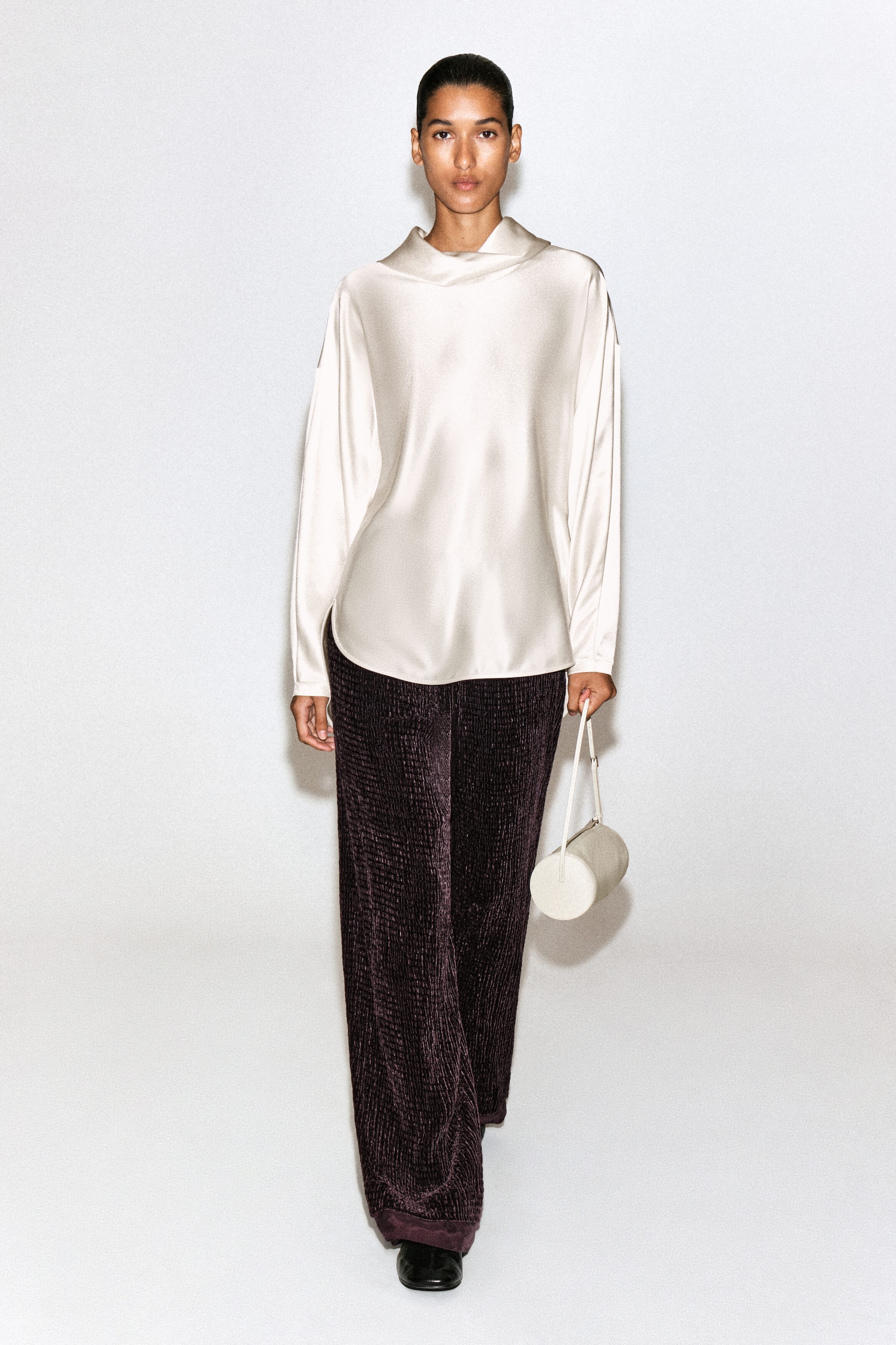 A champagne-colored satin long-sleeve top featuring a draped cowl neckline and a curved hem is shown from the front, falling loosely over dark purple, textured wide-leg pants. A champagne-colored satin long-sleeve top featuring a draped cowl neckline and a curved hem is shown from the front, falling loosely over dark purple, textured wide-leg pants.