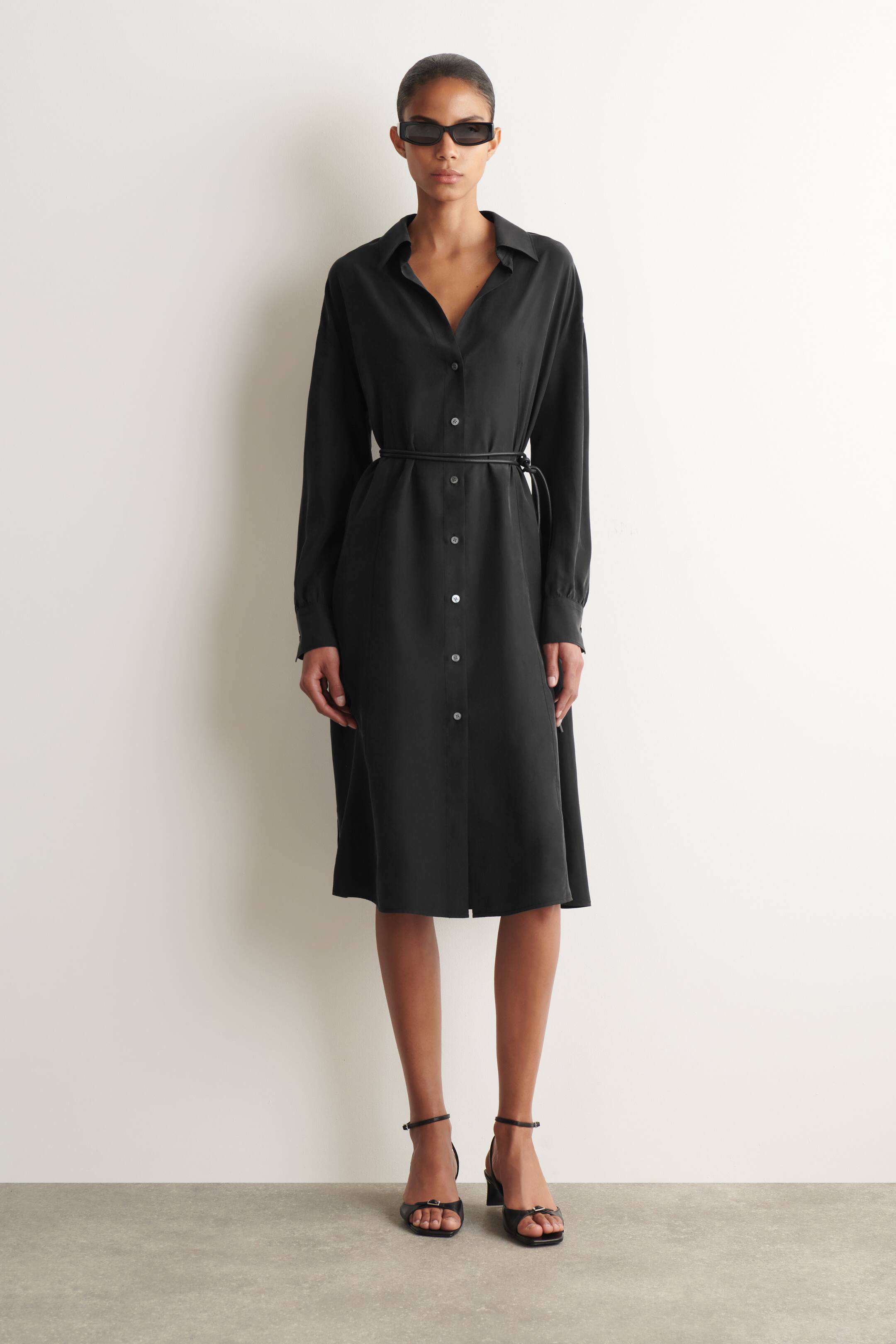 Agrandir l'image: Une robe-chemise midi noire présente un col pointu et des manches longues avec des poignets boutonnés. Elle est conçue avec une patte de boutonnage complète de boutons de couleur claire et est cintrée à la taille par une ceinture fine noire.