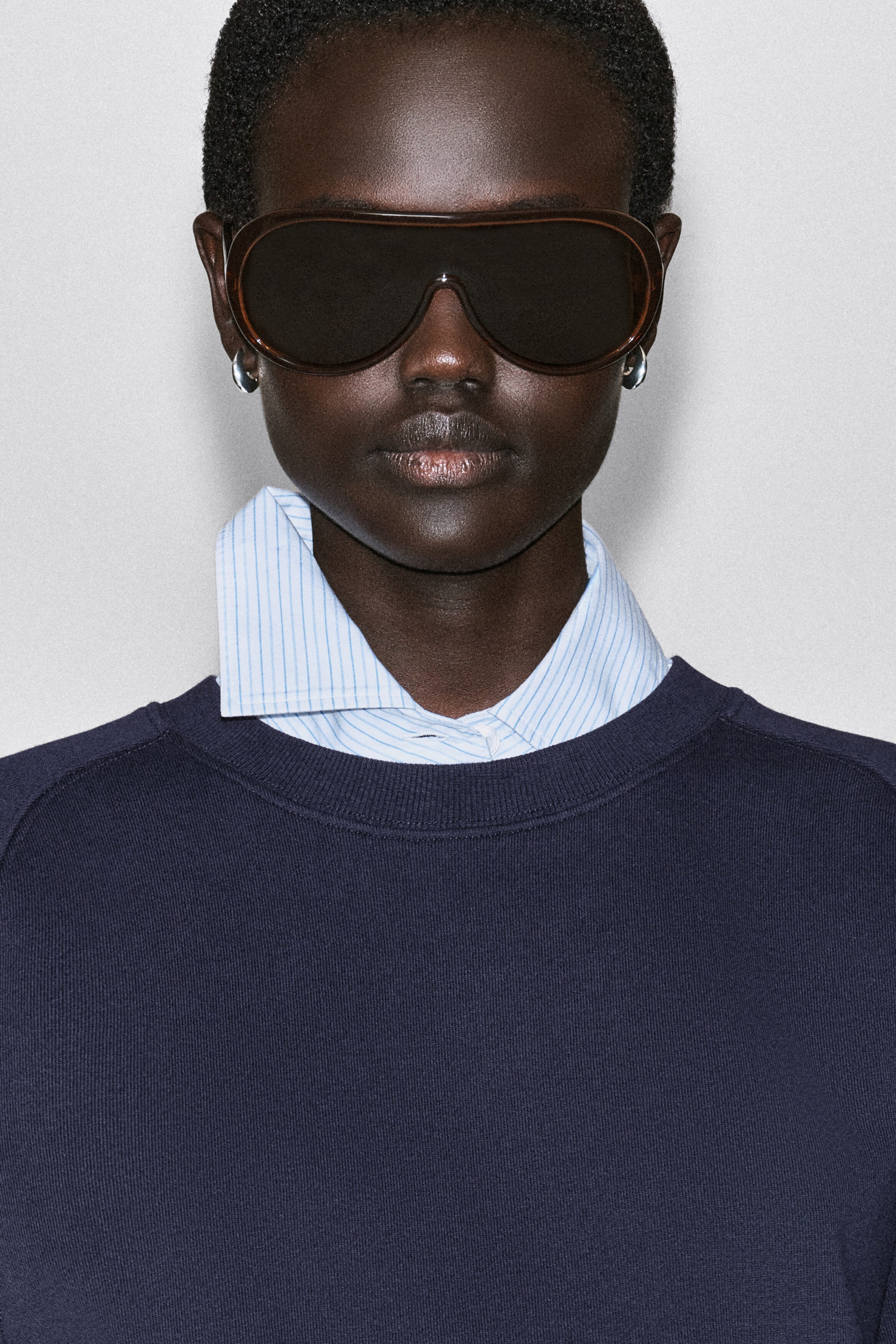 A navy blue cotton sweatshirt is being worn as a top, featuring a ribbed crew neck and raglan shoulder seams. The sweatshirt is layered over a light blue pinstriped collared shirt that is visible at the neckline. A navy blue cotton sweatshirt is being worn as a top, featuring a ribbed crew neck and raglan shoulder seams. The sweatshirt is layered over a light blue pinstriped collared shirt that is visible at the neckline.