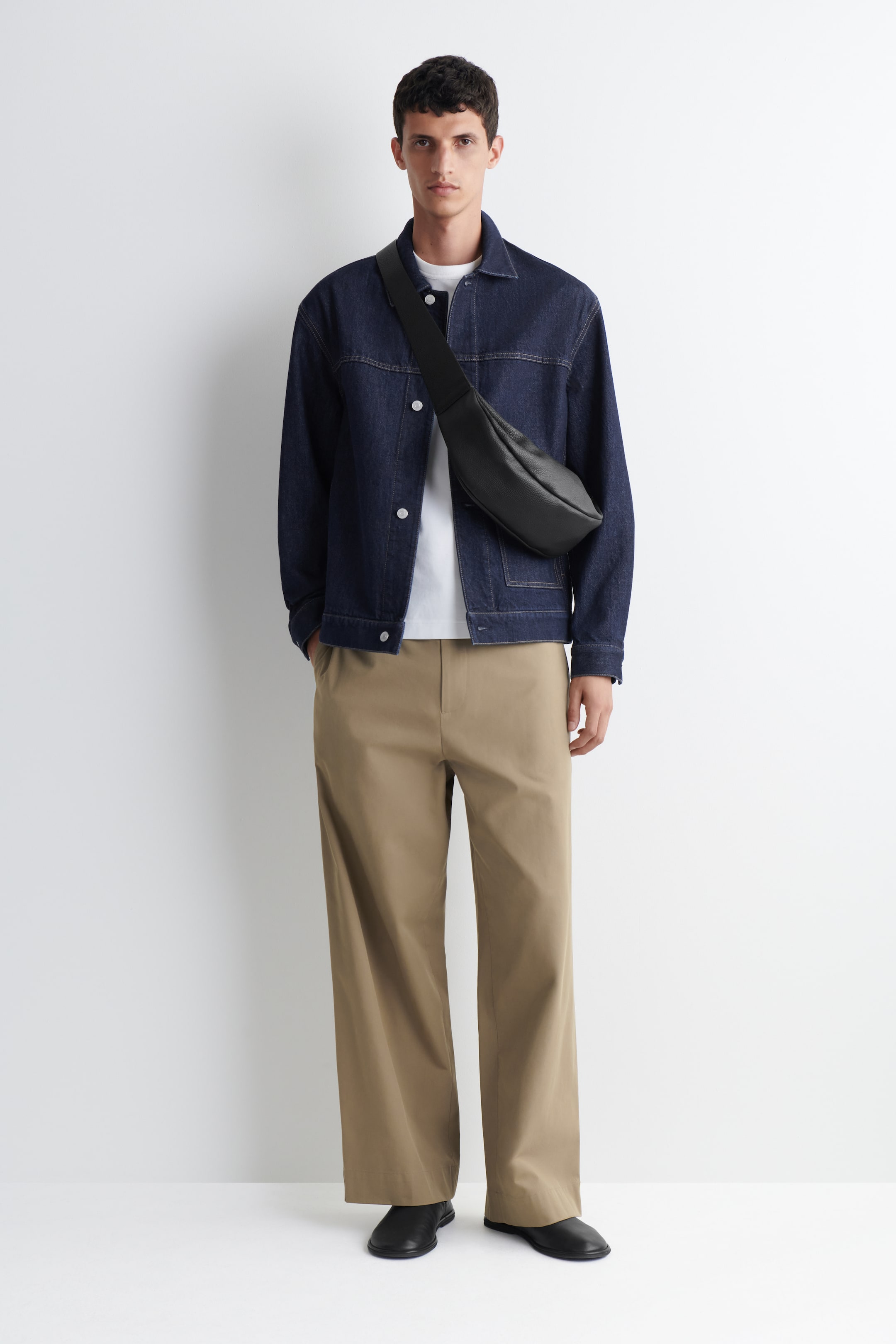 Agrandir l'image: Sac bandoulière noir, avec une large sangle, se courbant sur la poitrine et la hanche du mannequin, mettant en valeur son matériau texturé sur une veste en jean foncé.