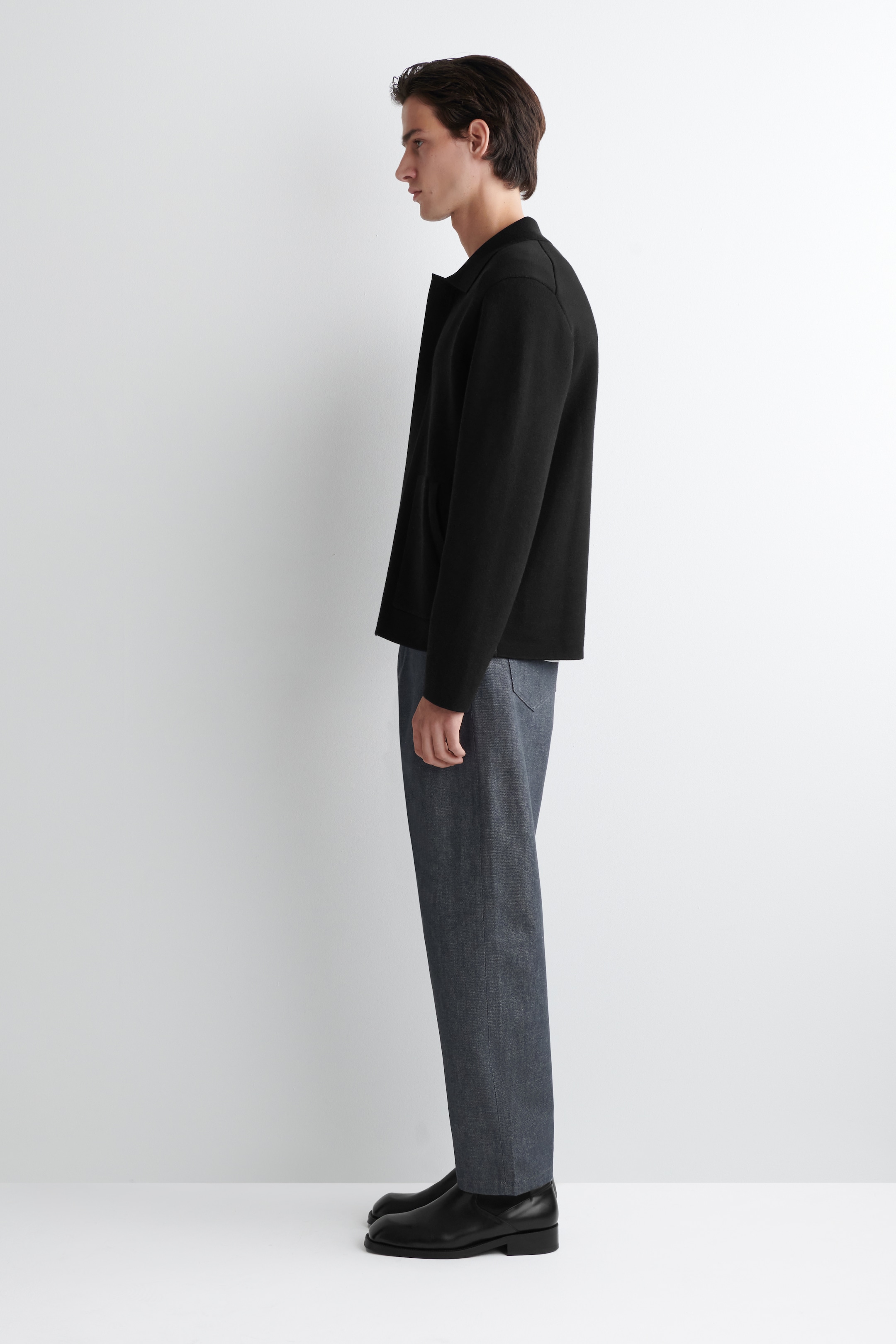 Agrandir l'image: Une veste noire, des pantalons gris et des chaussures noires sont modelés en profil complet de côté, face à droite. La veste noire présente un col évasé et des manches longues avec une coupe décontractée. Les pantalons gris montrent une coupe droite dans un tissu texturé, et les chaussures noires présentent un design bas.
