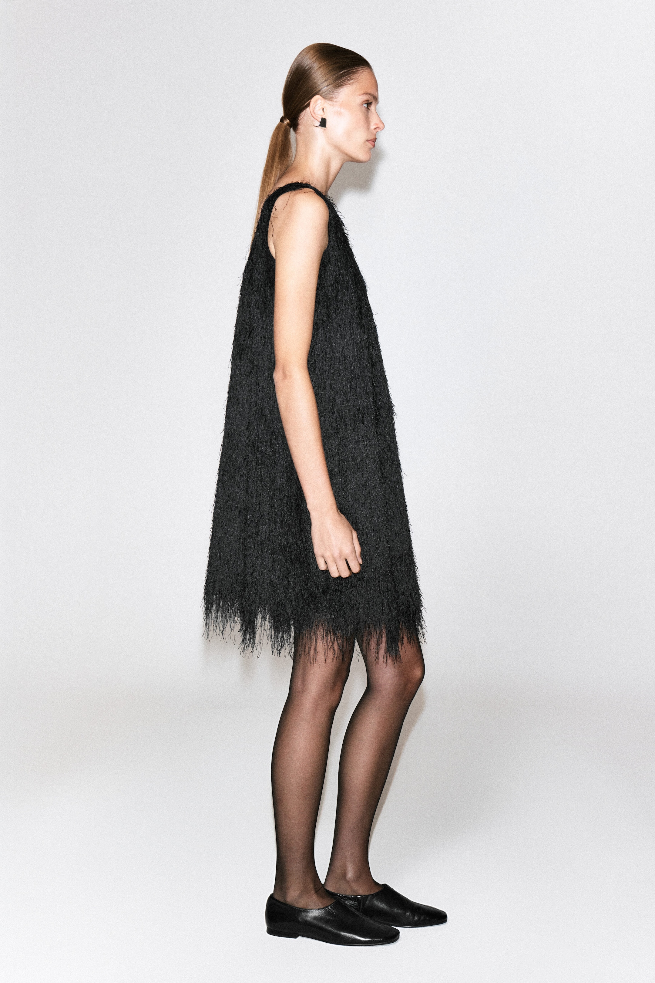 Ein schwarzes Minikleid mit Fransen wird von dem Model getragen. Es verfügt über eine ärmellose A-Linien-Silhouette, die vollständig mit feinen, strukturierten Fransen bedeckt ist. Das Kleid wird mit transparenten schwarzen Strumpfhosen und flachen schwarzen Schuhen kombiniert. Ein schwarzes Minikleid mit Fransen wird von dem Model getragen. Es verfügt über eine ärmellose A-Linien-Silhouette, die vollständig mit feinen, strukturierten Fransen bedeckt ist. Das Kleid wird mit transparenten schwarzen Strumpfhosen und flachen schwarzen Schuhen kombiniert.