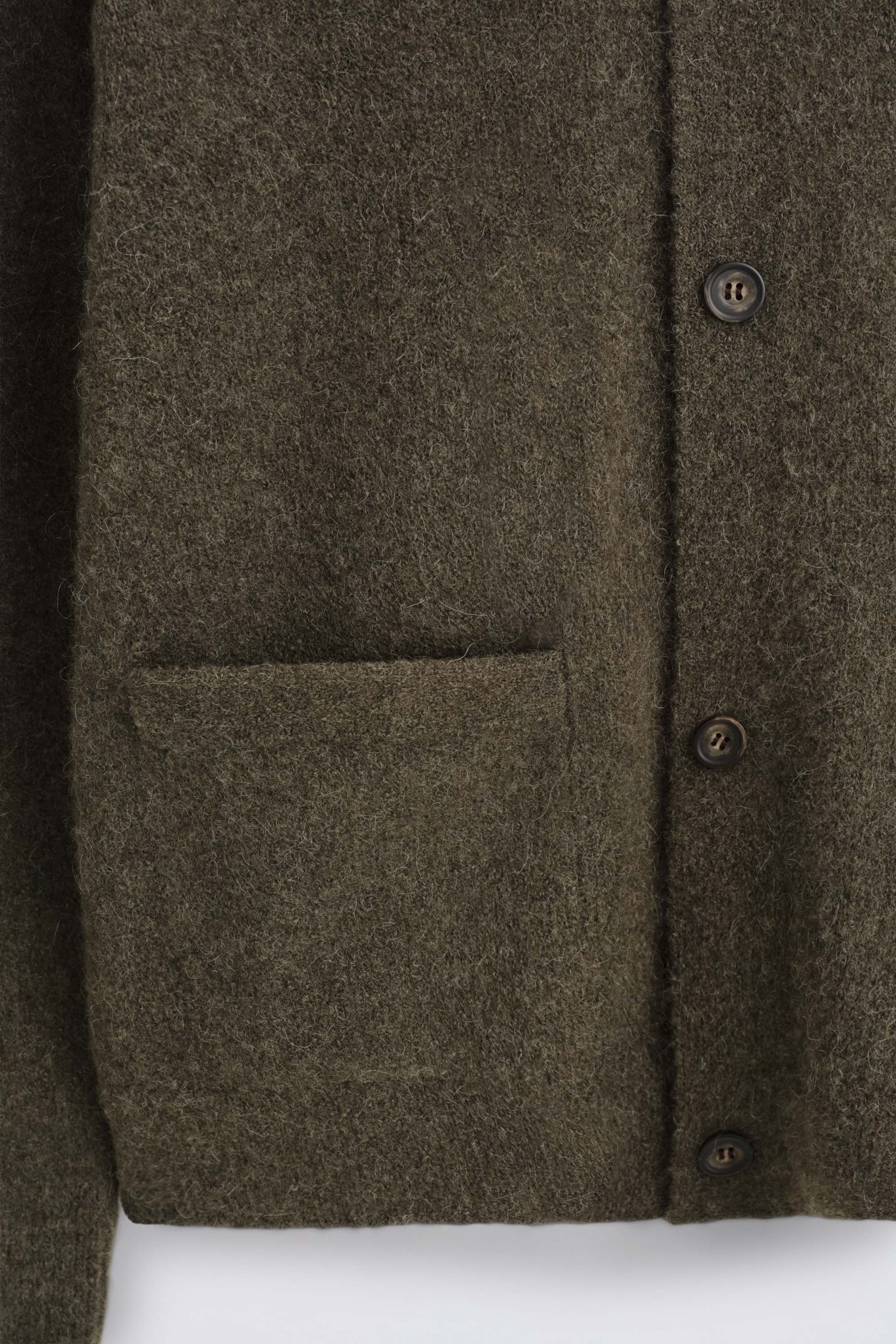 The textured collared cardigan is shown in a close-up view, featuring a fuzzy dark olive green wool-blend fabric. The front includes a flat patch pocket and a vertical button closure with dark round buttons. The textured collared cardigan is shown in a close-up view, featuring a fuzzy dark olive green wool-blend fabric. The front includes a flat patch pocket and a vertical button closure with dark round buttons.