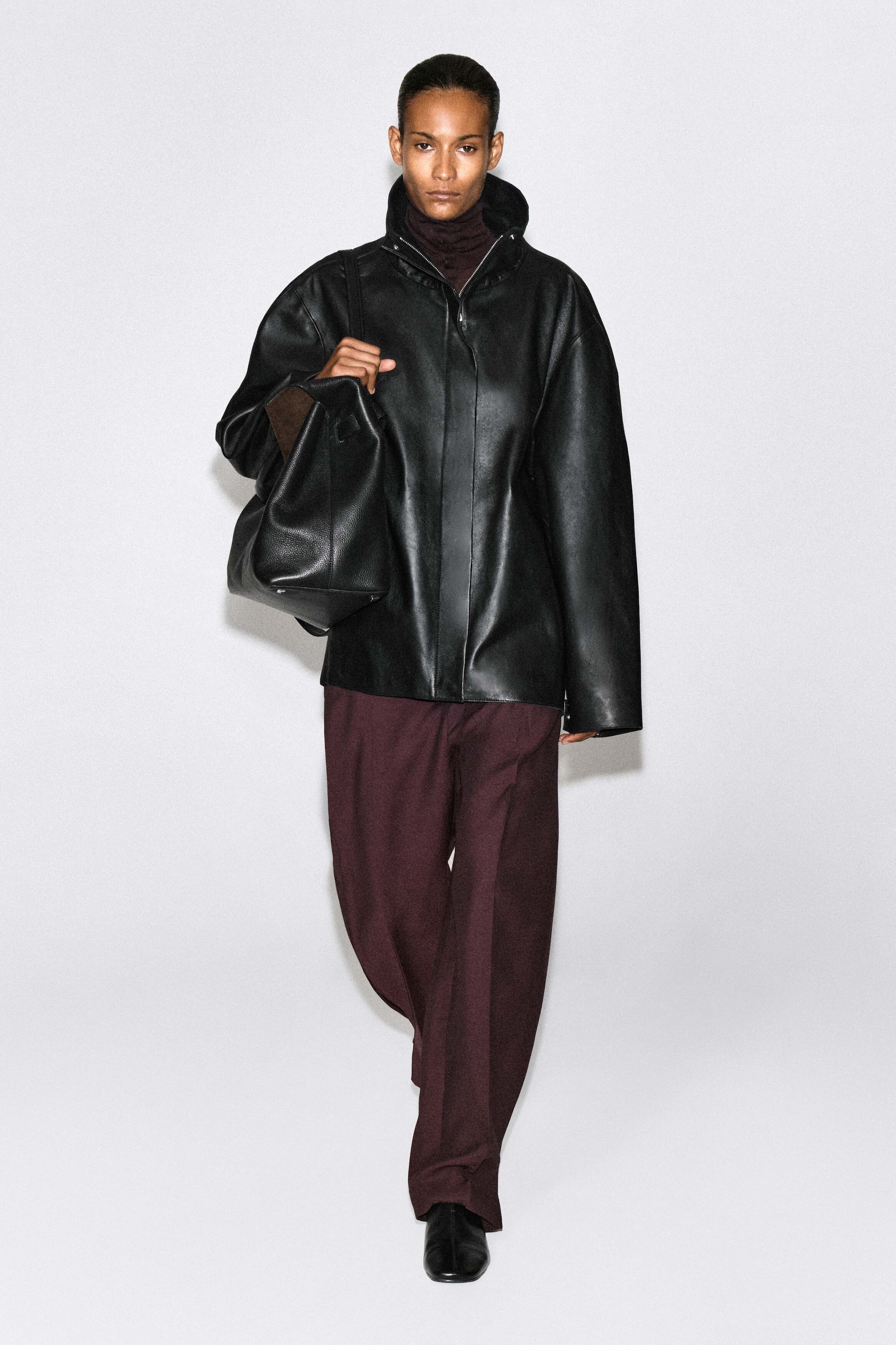 Pleated-panel straight-leg pants in deep burgundy are worn by the model walking forward. The relaxed-fit pants are styled with an oversized black leather jacket and a black leather bag draped over the shoulder. Pleated-panel straight-leg pants in deep burgundy are worn by the model walking forward. The relaxed-fit pants are styled with an oversized black leather jacket and a black leather bag draped over the shoulder.