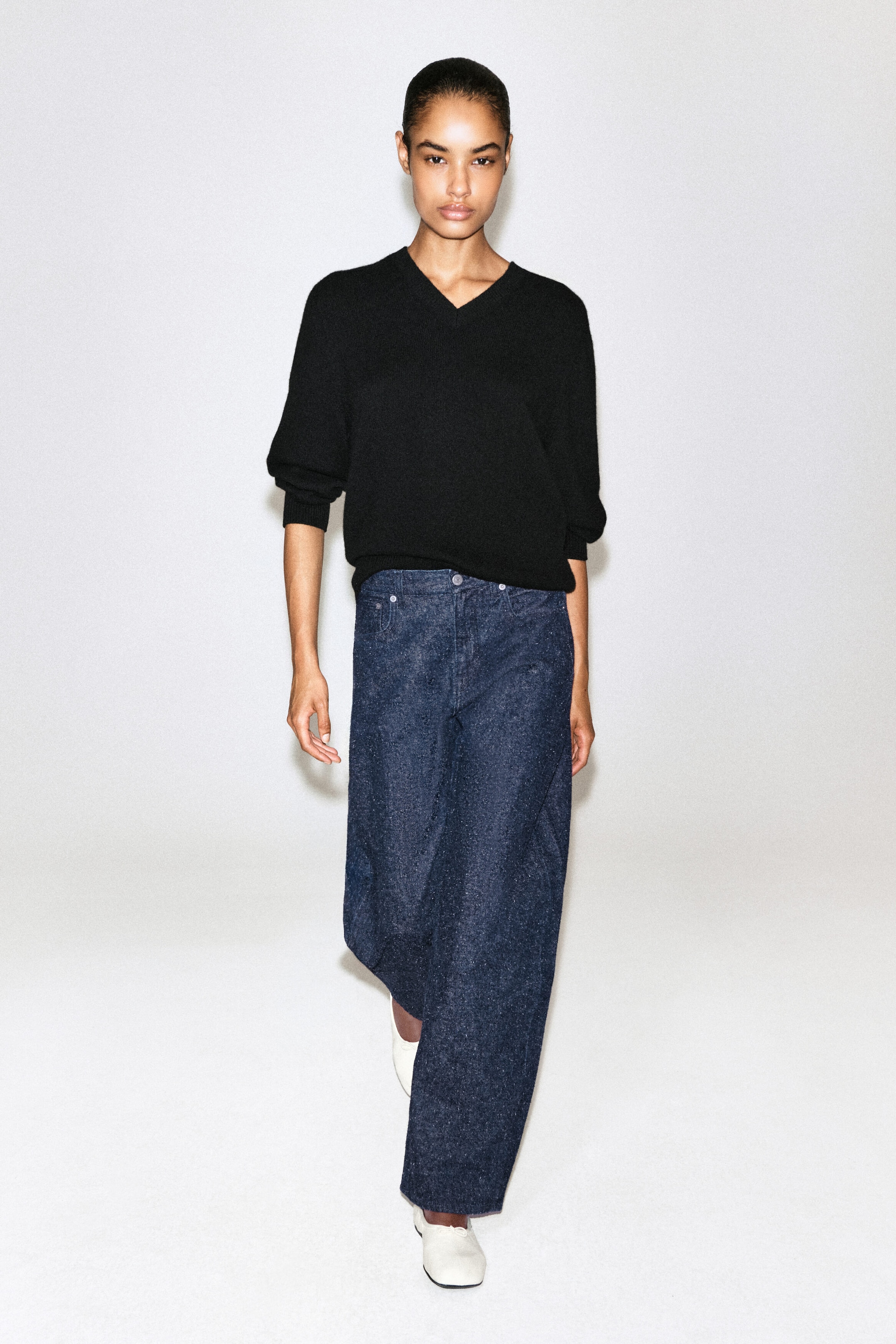 A model is presenting a black V-neck sweater and dark blue wide-leg jeans from the front. The relaxed-fit sweater shows pushed-up sleeves with ribbed cuffs, while the high-waisted, textured denim jeans drape into a wide, straight-leg silhouette. A model is presenting a black V-neck sweater and dark blue wide-leg jeans from the front. The relaxed-fit sweater shows pushed-up sleeves with ribbed cuffs, while the high-waisted, textured denim jeans drape into a wide, straight-leg silhouette.