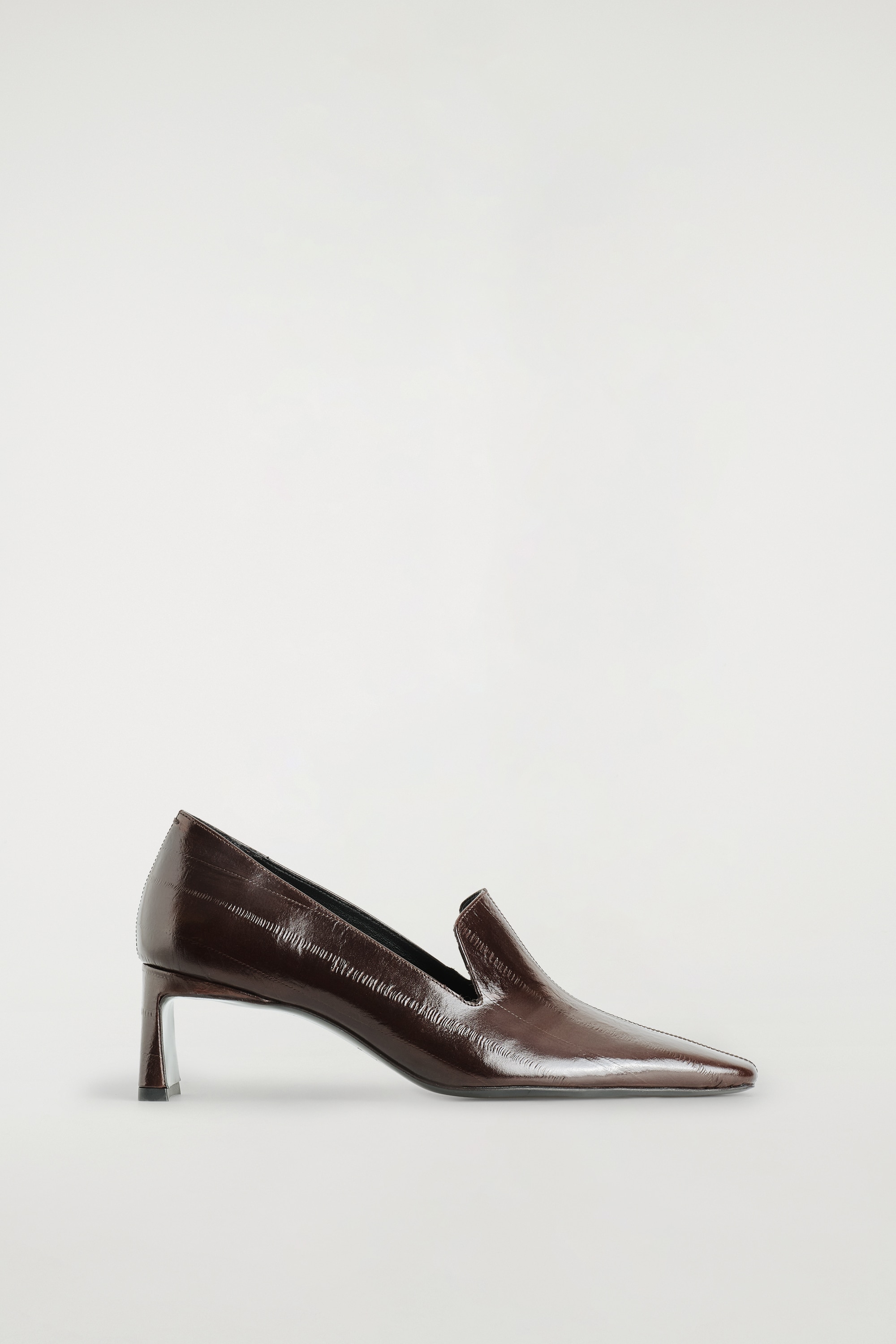 Sapatos loafers de salto em couro angular em castanho-escuro estÃ£o a ser usados de um Ã¢ngulo frontal, com calÃ§as escuras de perna larga a cair sobre os tornozelos. Os loafers apresentam uma biqueira angular distinta, de formato quadrado, e uma construÃ§Ã£o em couro liso.