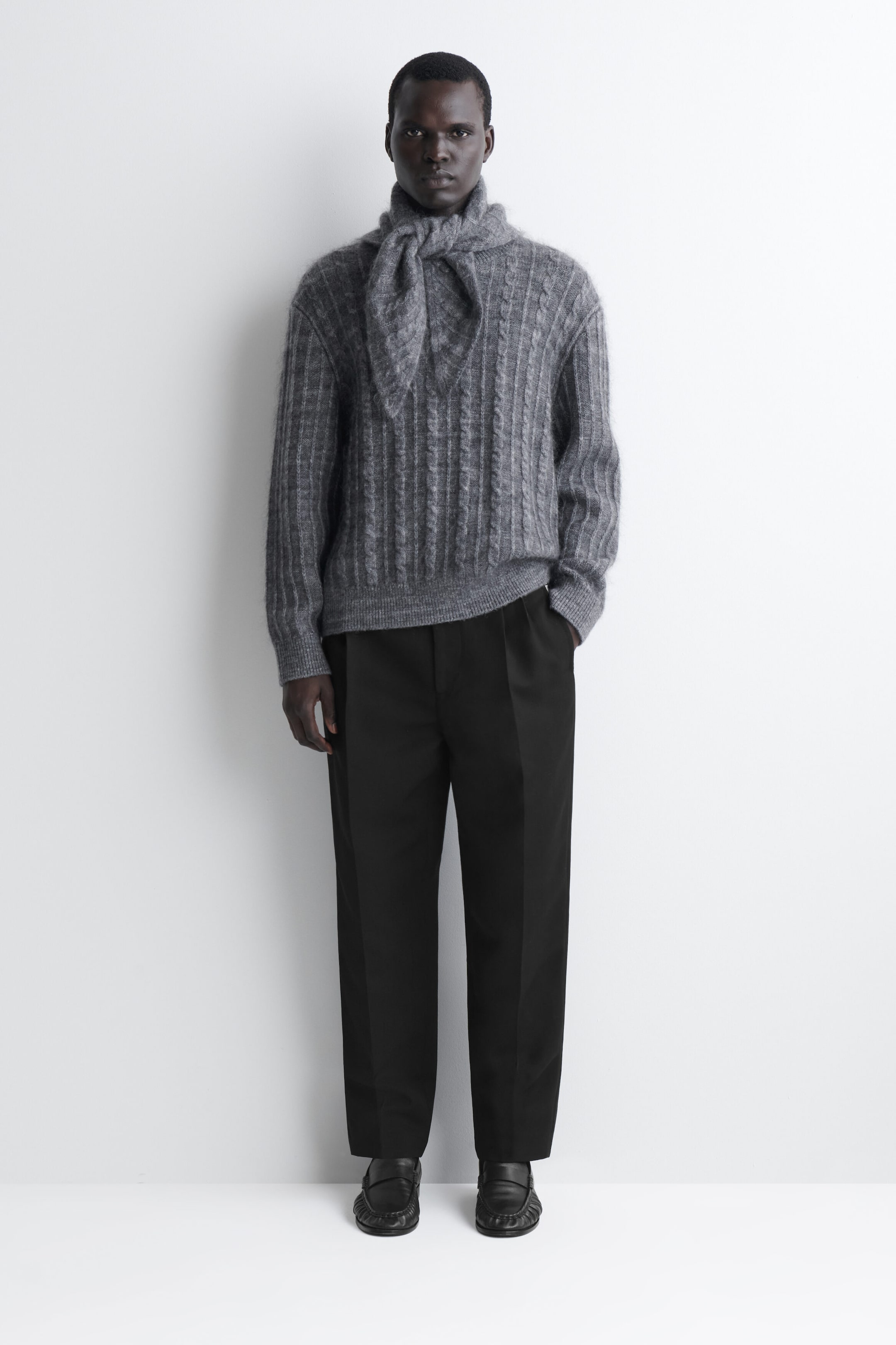 A grey cable-knit triangle scarf is being worn facing forward, styled with a loose knot at the neck over a matching textured sweater. The scarf showcases a soft, fuzzy knit texture and a triangular shape with pointed ends draped over the chest. A grey cable-knit triangle scarf is being worn facing forward, styled with a loose knot at the neck over a matching textured sweater. The scarf showcases a soft, fuzzy knit texture and a triangular shape with pointed ends draped over the chest.