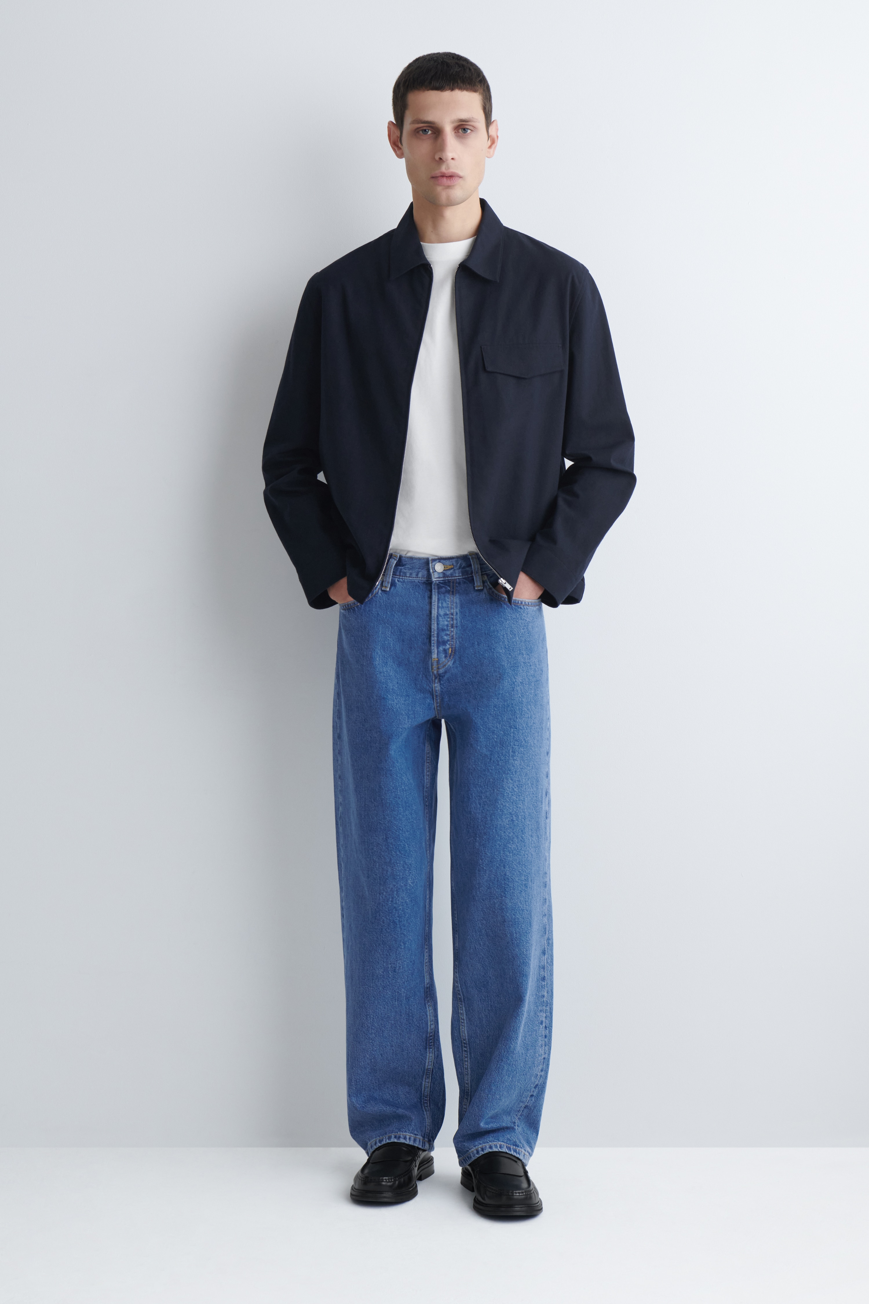 I jeans in denim blu sono presentati frontalmente, con una vestibilità relaxed e gamba dritta. Presentano una chiusura con bottoni a vista, tasche frontali a scoop con rivetti e un piccolo taschino portamonete sul lato destro.