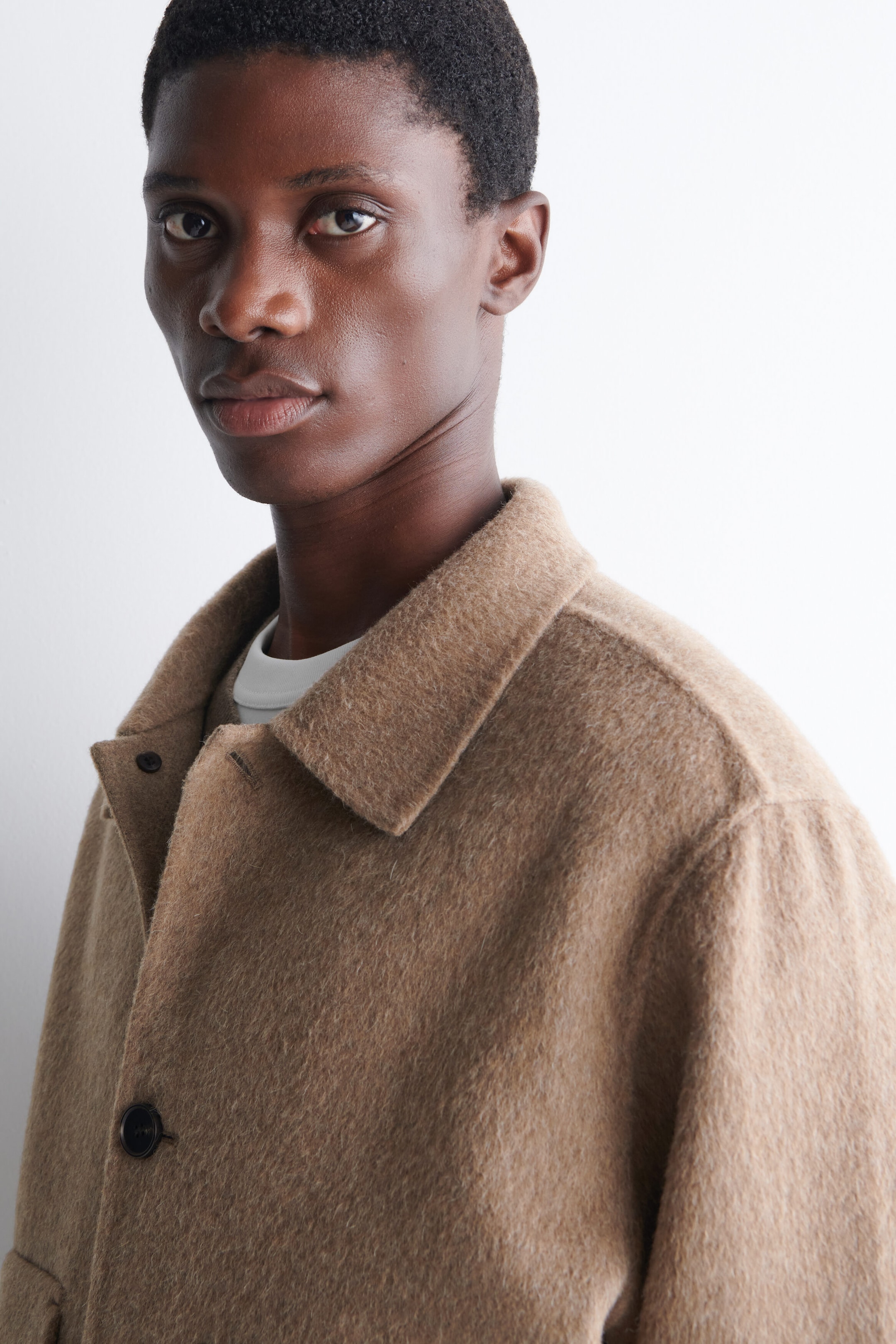 The model is wearing the double-faced blouson jacket in a textured light brown wool fabric. The jacket features a pointed collar and dark buttons and is layered over a white crewneck T-shirt. The model is wearing the double-faced blouson jacket in a textured light brown wool fabric. The jacket features a pointed collar and dark buttons and is layered over a white crewneck T-shirt.