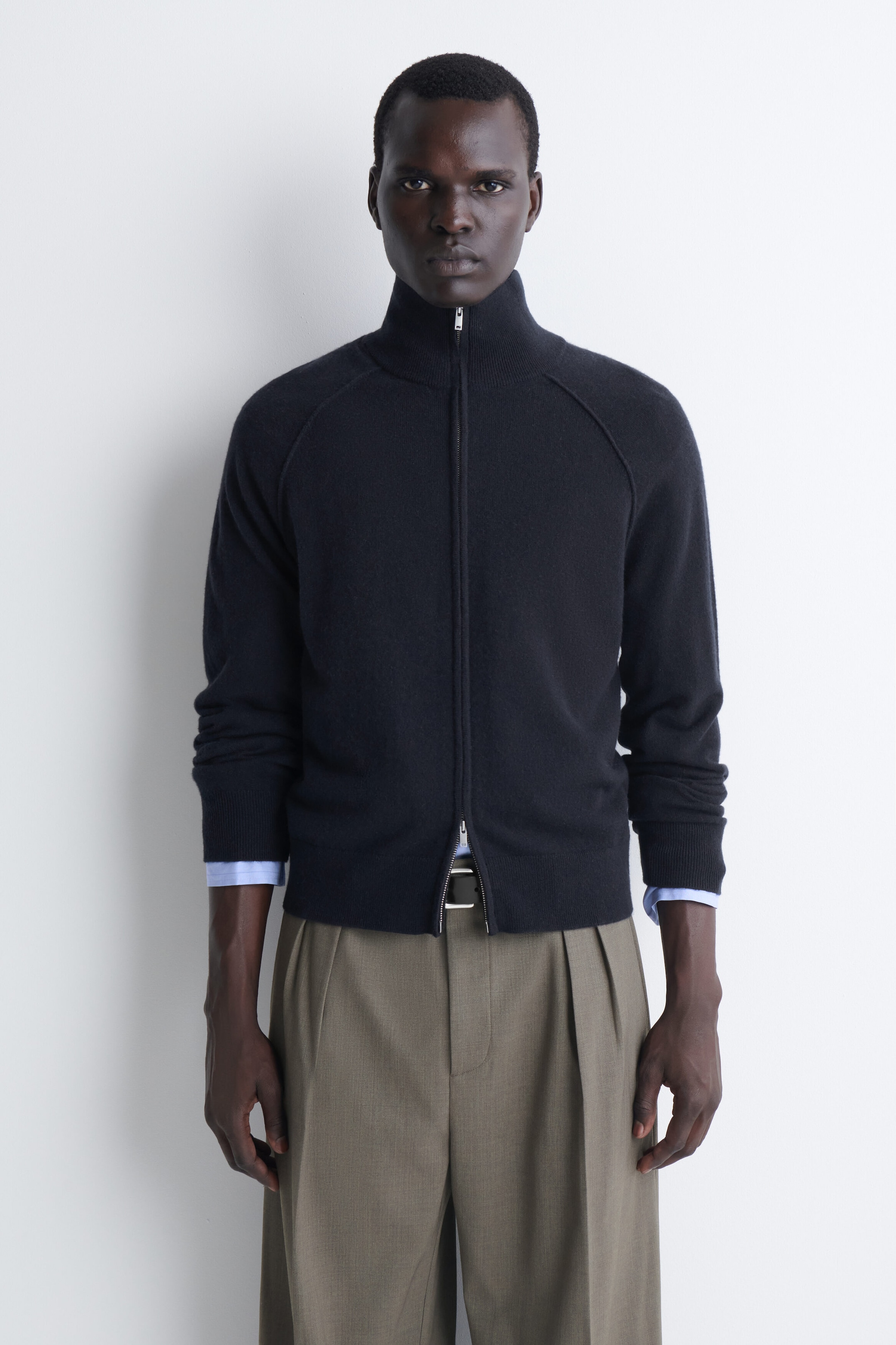 This front view showcases a dark navy cashmere funnel-neck zip-up cardigan featuring a two-way zip and raglan sleeves. The cardigan is styled zipped up to the neck, with the lower zip slightly open, and a light blue collared shirt peeking out from beneath the ribbed cuffs. This front view showcases a dark navy cashmere funnel-neck zip-up cardigan featuring a two-way zip and raglan sleeves. The cardigan is styled zipped up to the neck, with the lower zip slightly open, and a light blue collared shirt peeking out from beneath the ribbed cuffs.
