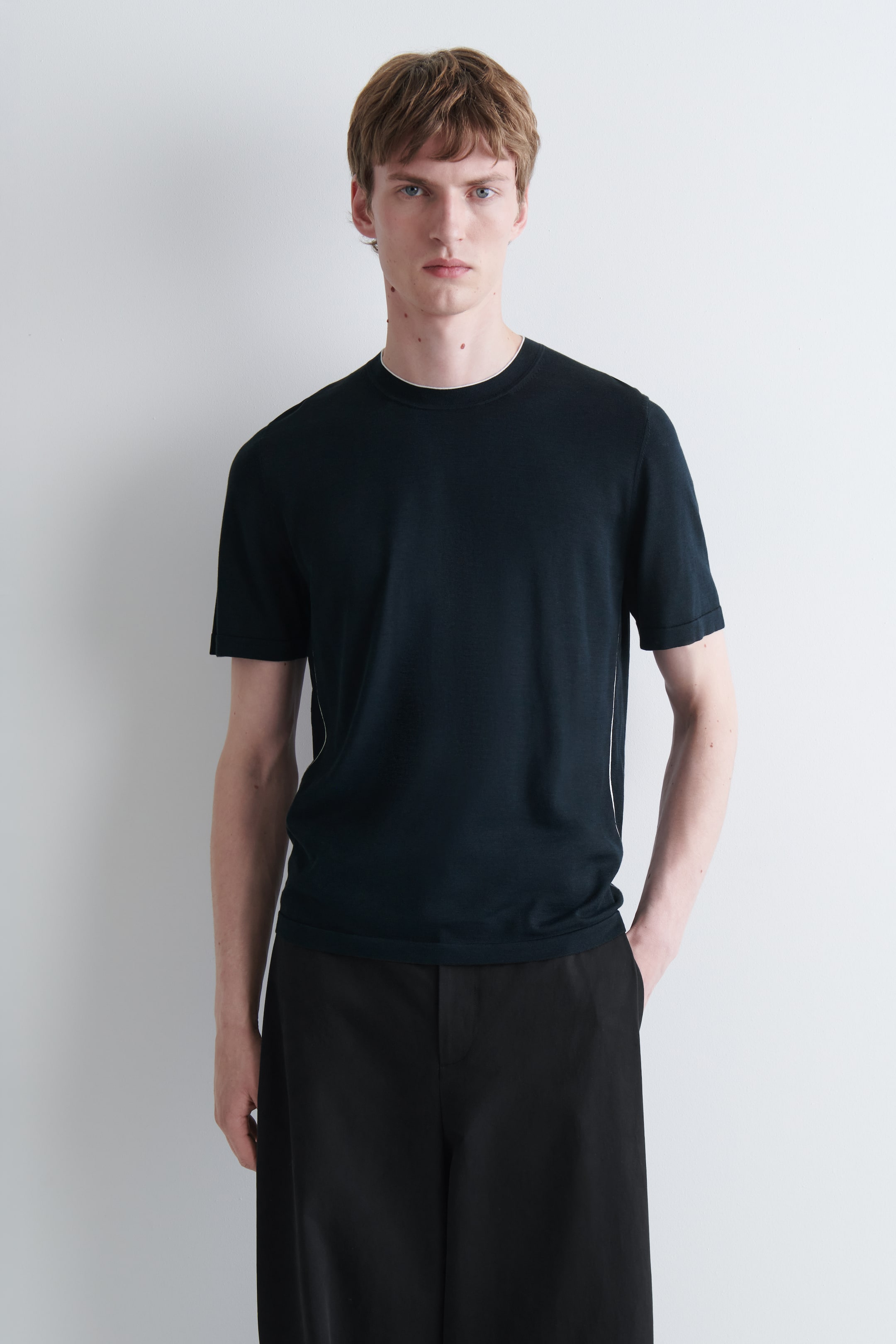 Agrandir l'image: Un T-shirt en maille sombre, à la coupe slim, à manches courtes, avec un passepoil contrastant blanc autour du col rond et le long des coutures latérales, est porté par le mannequin.