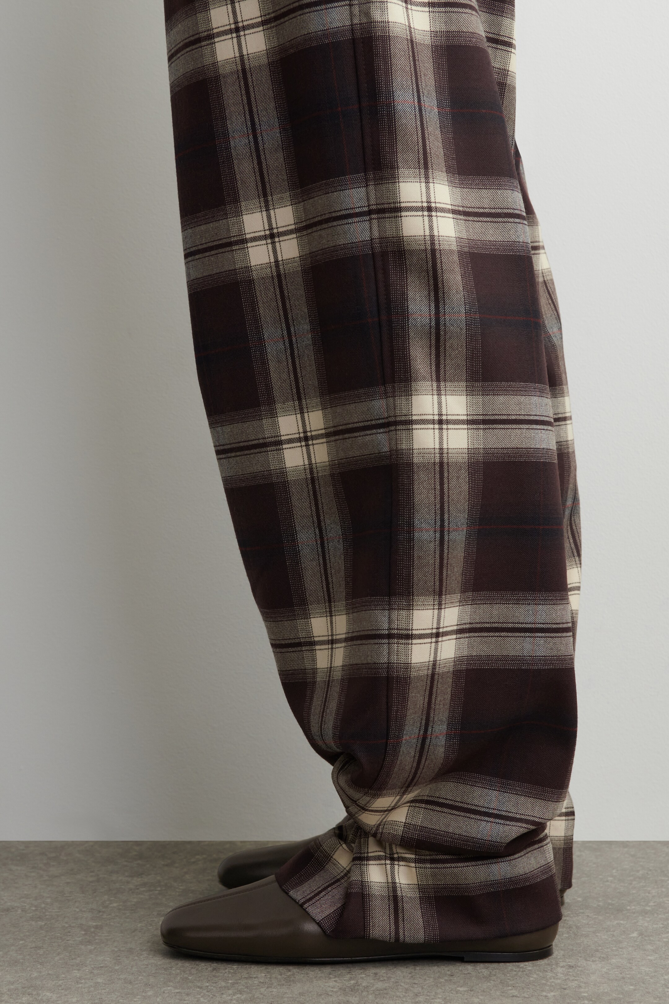 Agrandir l'image: Un pantalon est présenté de côté, avec un motif à carreaux marron foncé et crème, avec des lignes subtiles grises, rouges et bleues. Le pantalon ample est regroupé sur des chaussures plates marron foncé, mettant en valeur leur ourlet pleine longueur.