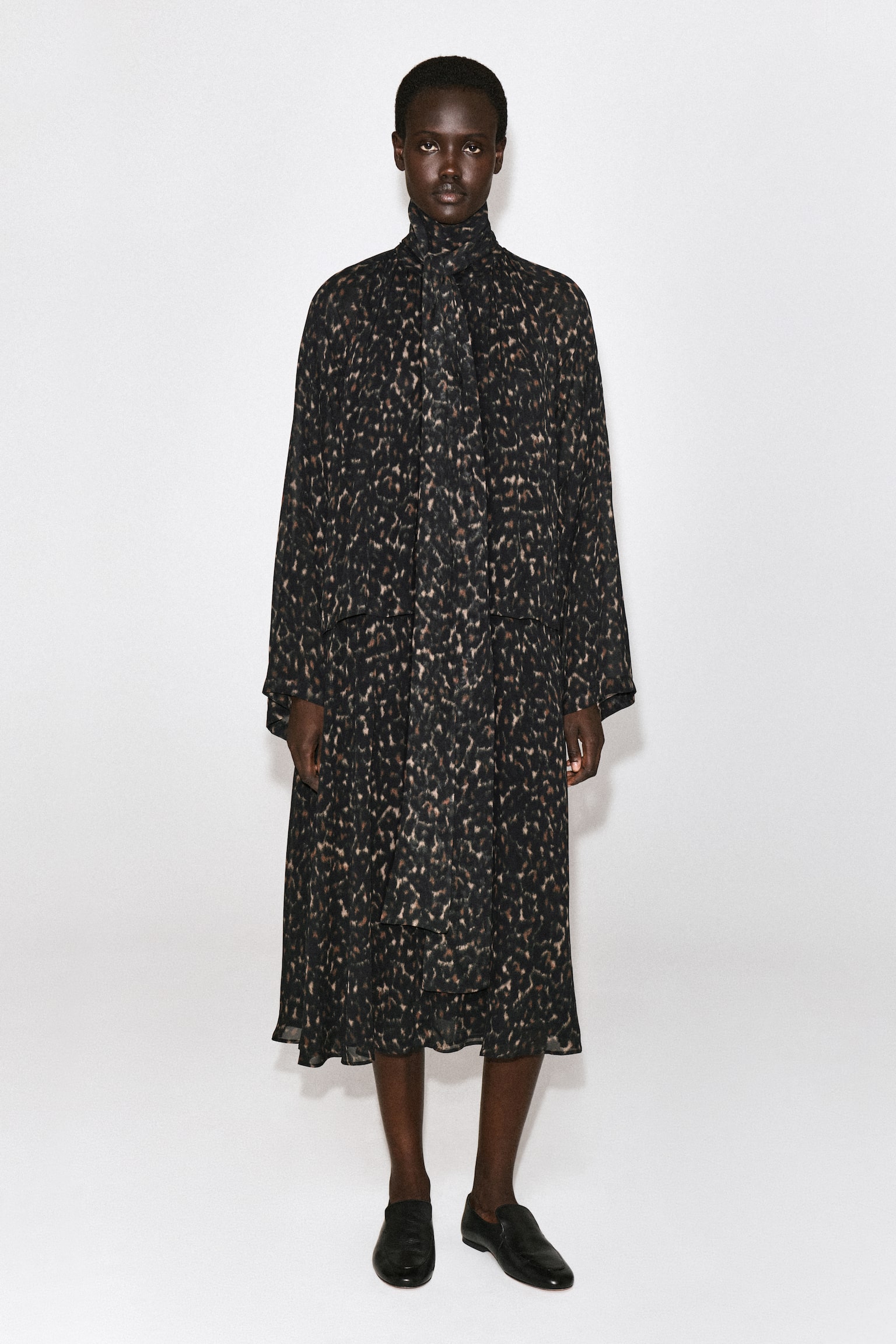 O vestido fluido com estampado de leopardo e detalhe de lenço está pendurado, visto de frente, exibindo uma gola alta com detalhe de lenço amovÃvel atado ao pescoço. Apresenta mangas compridas e largas, um corpete de design em camadas e uma silhueta descontraÃda e fluida, com padrão integral de leopardo em castanho e preto. - 1
