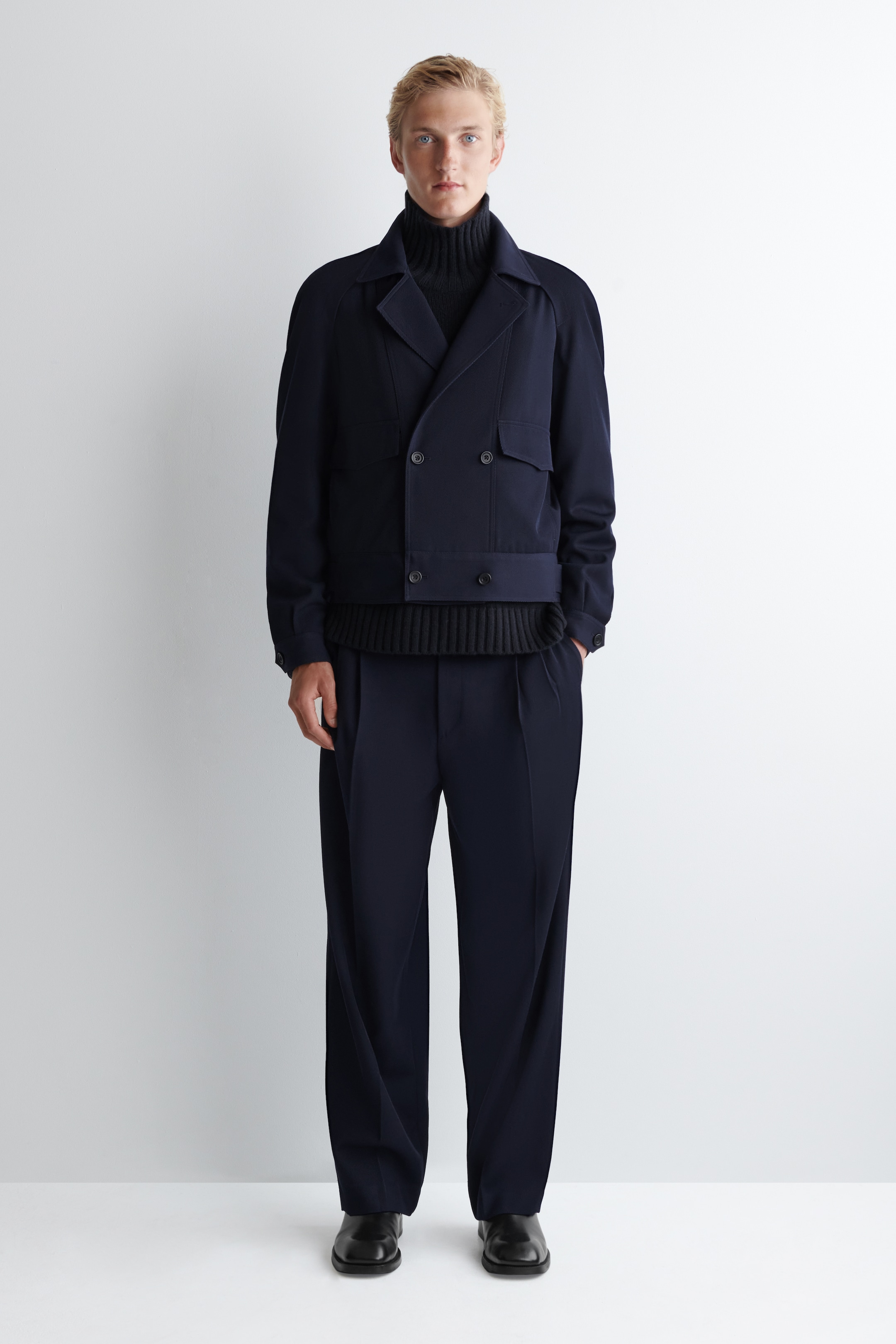 View larger image: A front-facing dark navy ensemble is being presented, featuring a double-breasted jacket with two flap pockets and dark buttons, layered over a black ribbed turtleneck sweater with a visible ribbed hem. Matching wide-leg pleated trousers are completing the look, pooling over black dress shoes.