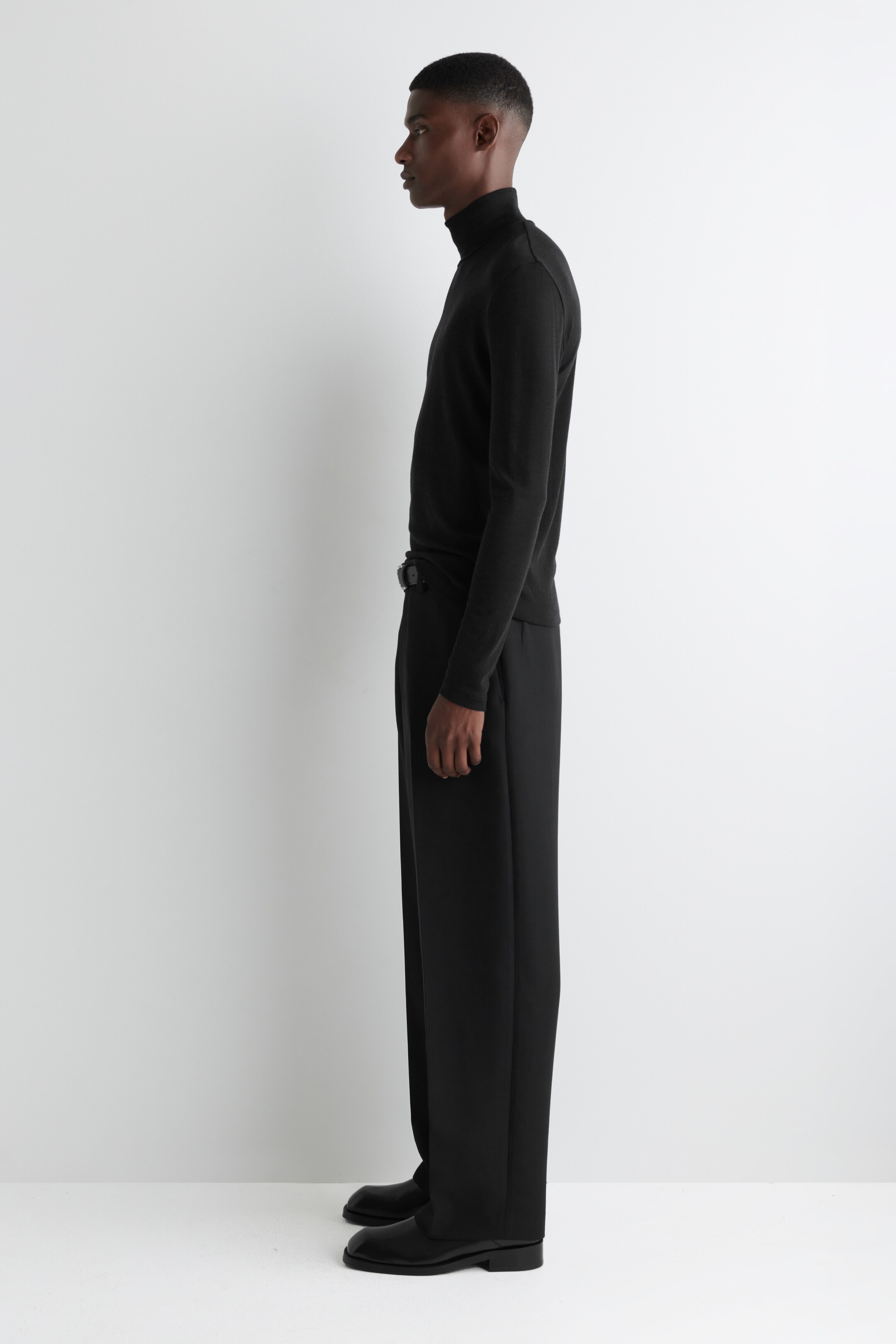 View larger image: A black long-sleeved turtleneck top is being worn, showing its slim fit and high neck, presented from a side profile. It is being paired with loose-fitting black wide-leg trousers, which are cinched by a dark belt, and black shoes.