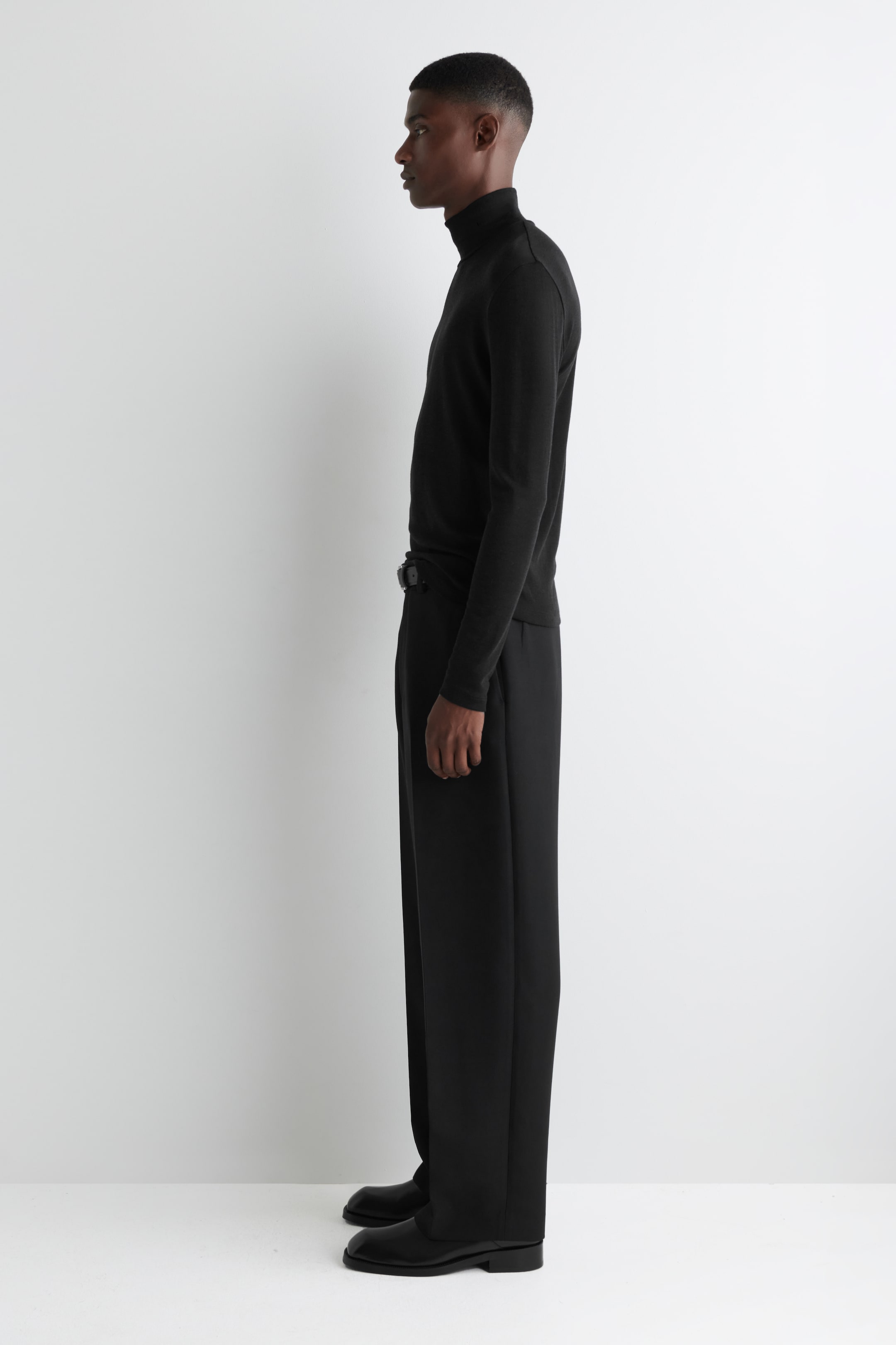 Agrandir l'image: Un haut à col roulé noir à manches longues est porté, montrant sa coupe ajustée et son col haut, présenté de profil. Il est associé à un pantalon noir à jambes larges et ample, serré par une ceinture foncée, et des chaussures noires.