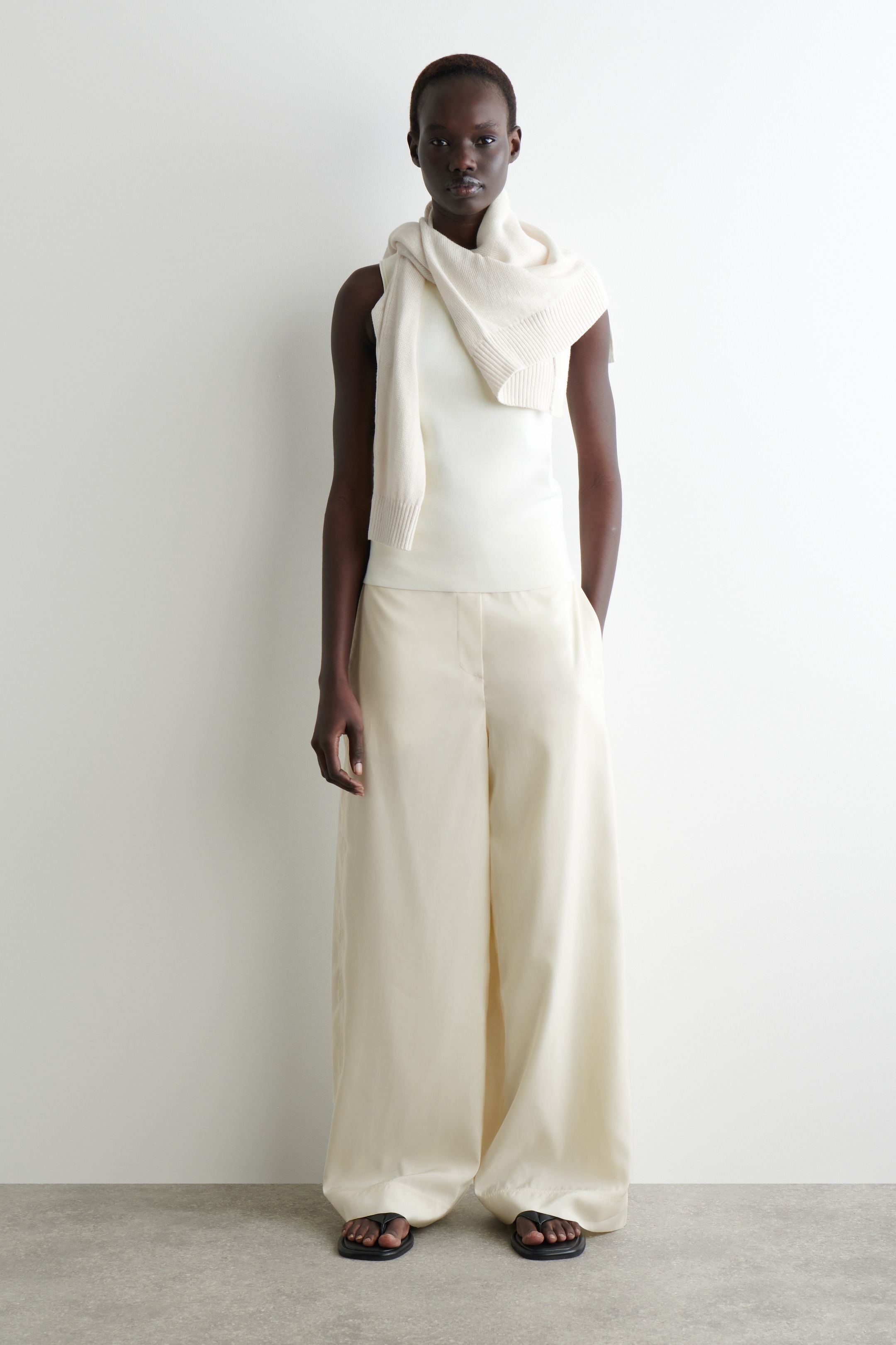 Agrandir l'image: Une tenue monochrome blanc cassé est présentée, avec un haut sans manches superposé sous une écharpe en tricot drapée autour du cou et des épaules, montrant des détails côtelés. Un pantalon à jambes larges dans une teinte blanc cassé assortie est porté, exhibant un drapé lisse et fluide, et des sandales noires à entre-doigts complètent le look.