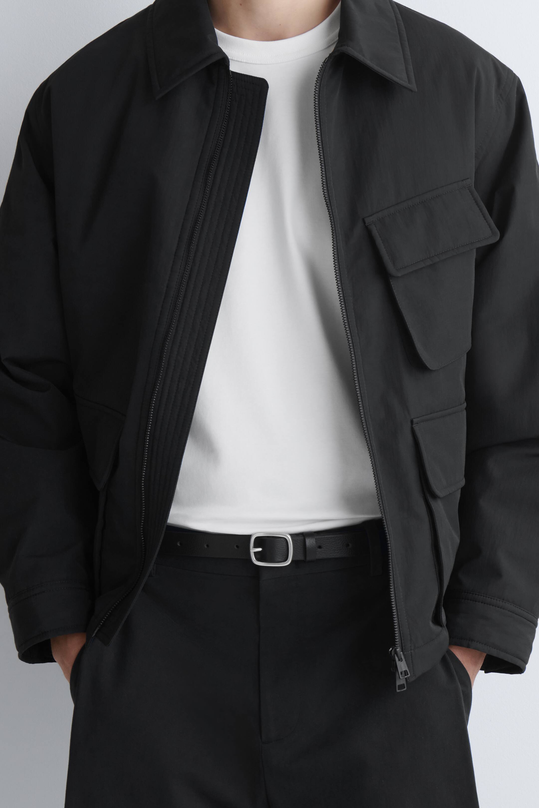 Grösseres Bild ansehen: Eine schwarze Jacke wird von vorne präsentiert, teilweise geöffnet, sodass ein weißes T-Shirt darunter sichtbar wird. Sie verfügt über einen spitzen Kragen, zwei Pattentaschen auf der rechten Vorderseite und ein geripptes Panel entlang der inneren Reißverschlussleiste.