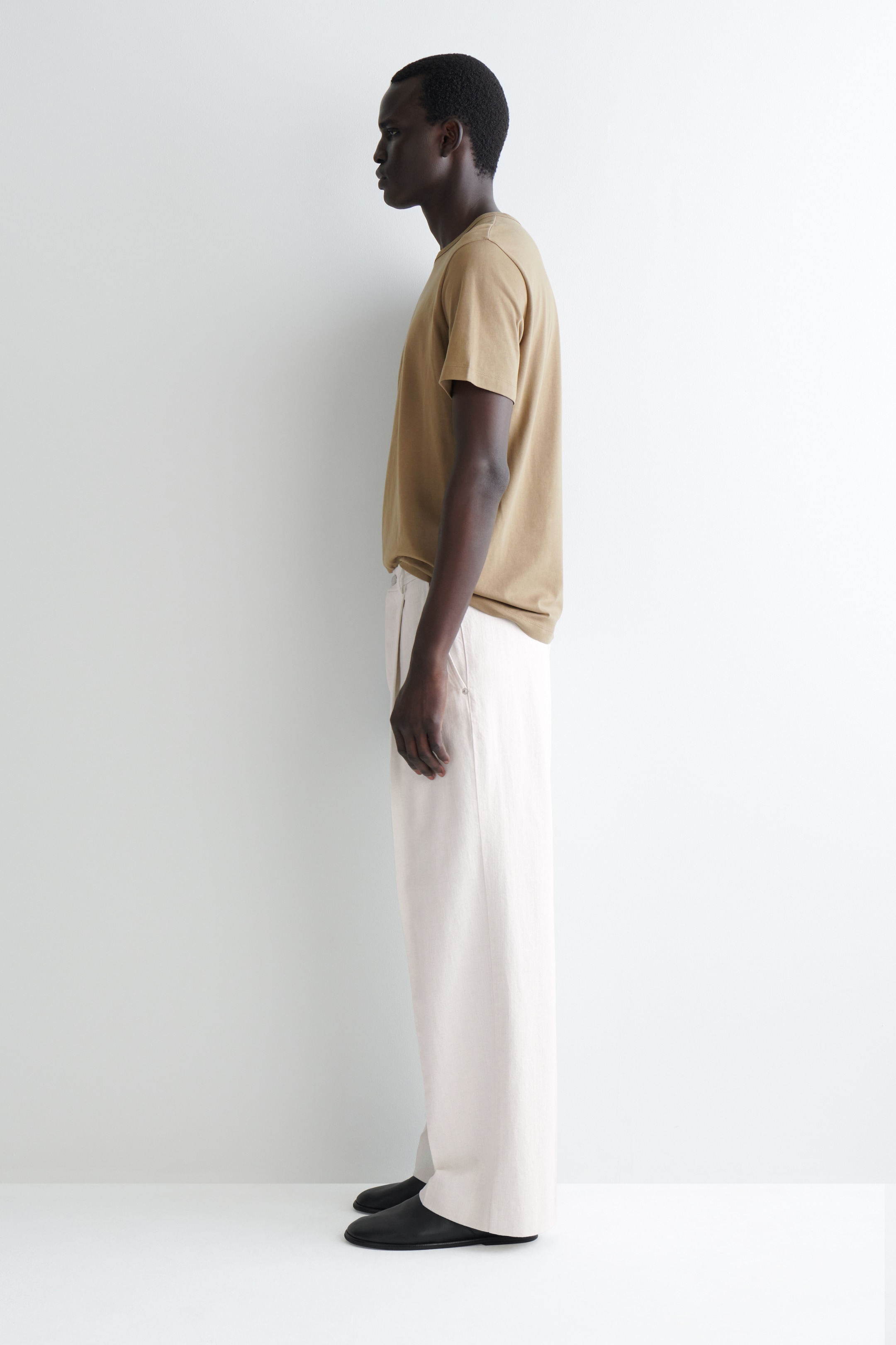 Agrandir l'image: Un t-shirt à col rond marron clair est porté, ses manches courtes et sa coupe décontractée sont visibles de profil. Un pantalon crème à jambes larges complète le t-shirt, tombant librement et couvrant des chaussures plates noires.