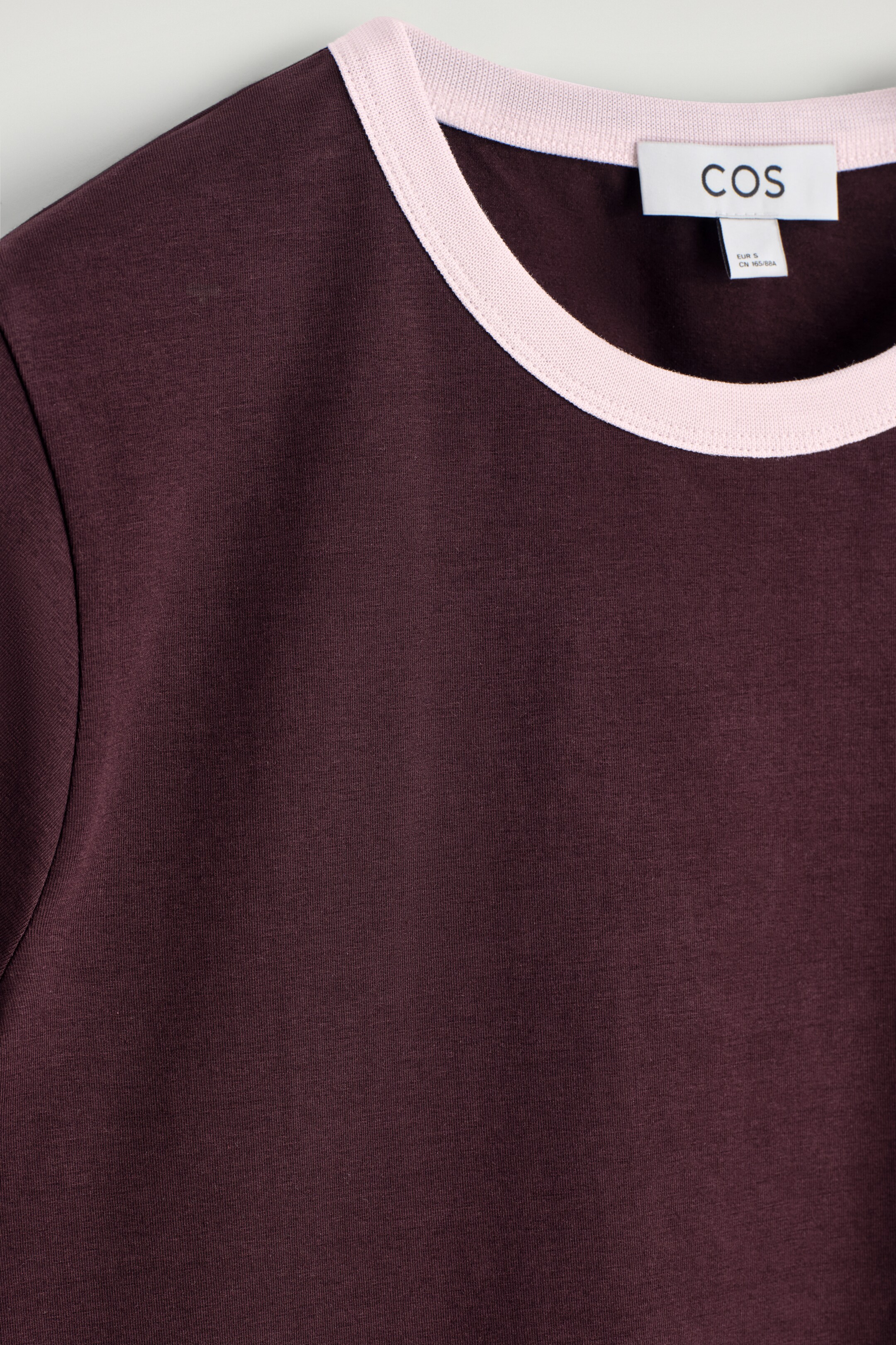 Vezi imaginea mai mare: Un tricou maroon cu ringer este prezentat, arătând partea din față. Acesta are un guler rotund cu bordură contrastantă roz pal. O etichetă albă de brand COS este vizibilă pe partea interioară a gâtului la spate.