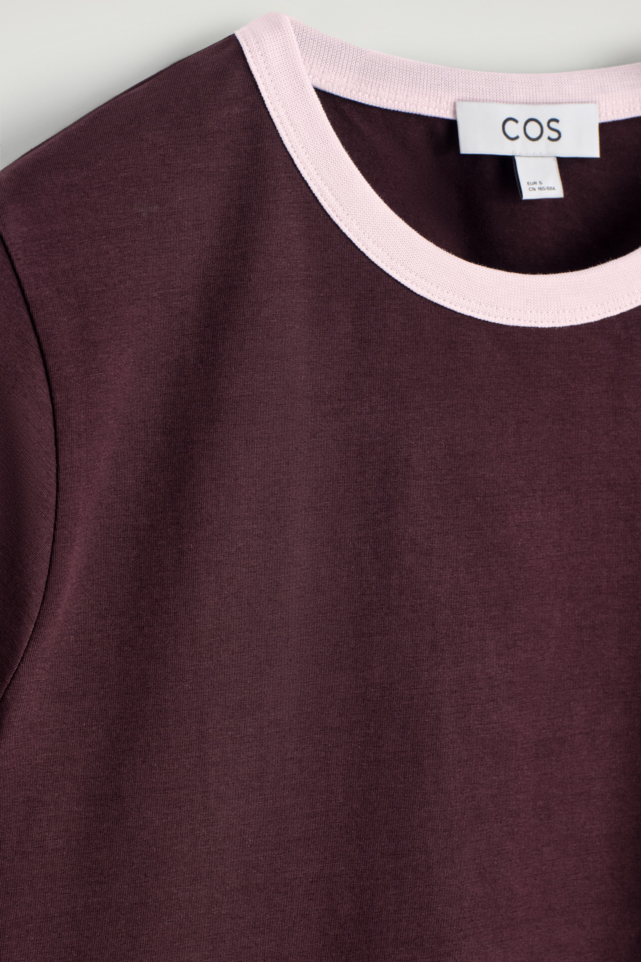 Agrandir l'image: Un T-shirt à col rond bordeaux est présenté, montrant son devant. Il a un col ras du cou avec un passepoil contrastant rose pâle. Une étiquette de marque COS blanche est visible à l'intérieur du col arrière.
