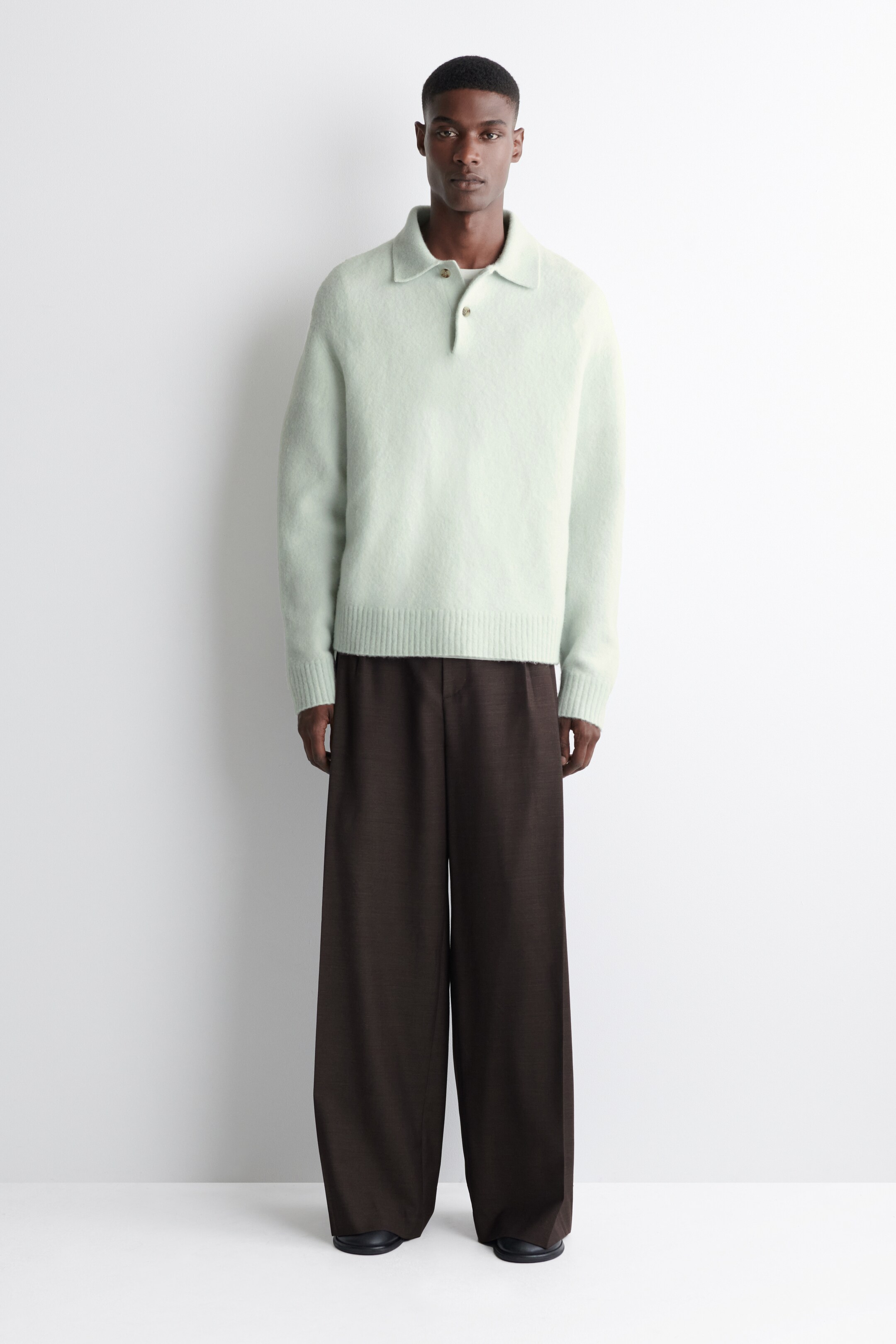 Visualizza immagine più grande: Un maglione polo a maniche lunghe verde chiaro presenta una texture morbida e soffice, un'abbottonatura a due bottoni e polsini e orlo a coste, rivolto in avanti. È abbinato a pantaloni marrone scuro a gamba larga che cadono morbidi.