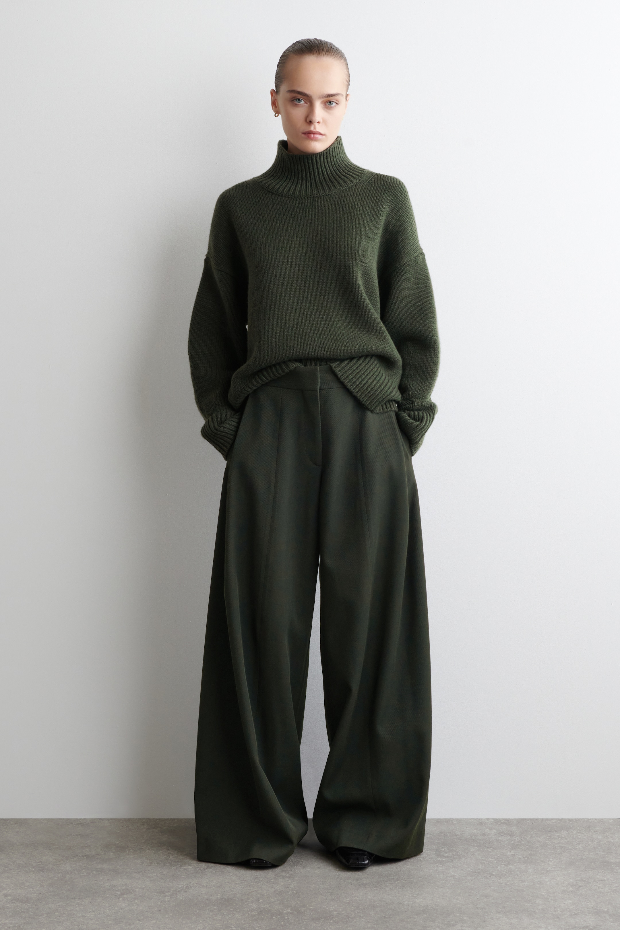 Visualizza immagine più grande: Un maglione a collo alto verde scuro, a vestibilità rilassata, con dettagli a coste e polsini ripiegati è abbinato a pantaloni a vita alta e gamba larga coordinati, caratterizzati da una piega centrale sul davanti. L'orlo anteriore del maglione appare leggermente infilato nella cintura, e i pantaloni si accumulano sopra le calzature.