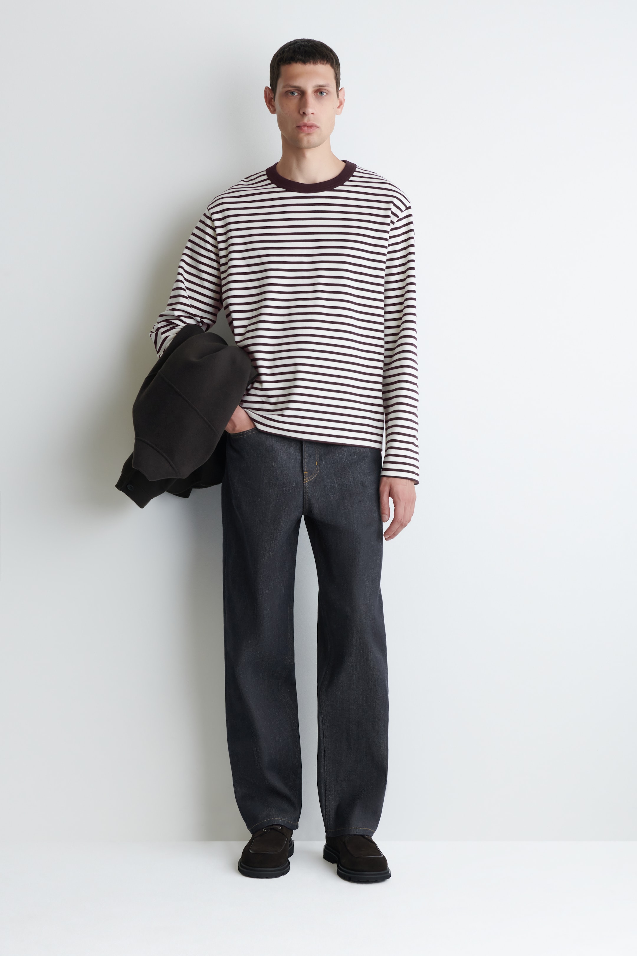 Agrandir l'image: Une chemise à manches longues, blanc cassé avec des rayures horizontales foncées et un col rond côtelé bordeaux est portée. Un jean à large jambe, de couleur foncée avec des coutures contrastantes orange est porté, accompagné de mocassins en daim foncé avec des semelles épaisses.