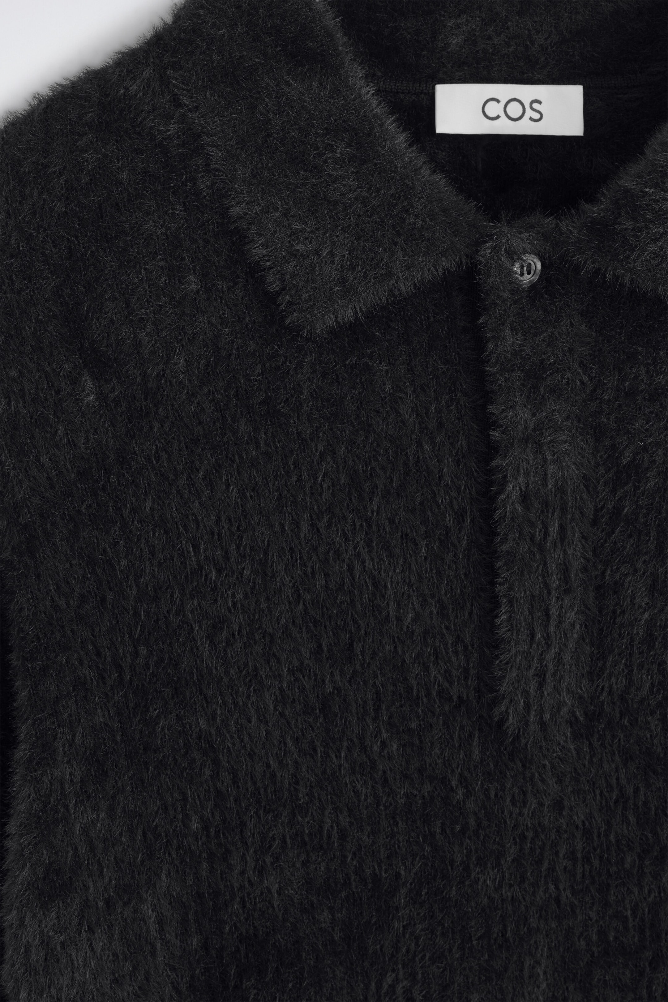 A close-up front view shows a black knitted polo shirt with a highly textured, fuzzy brushed finish. The shirt features a pointed collar, a button placket with one visible button, and a white brand label on the inside collar. A close-up front view shows a black knitted polo shirt with a highly textured, fuzzy brushed finish. The shirt features a pointed collar, a button placket with one visible button, and a white brand label on the inside collar.