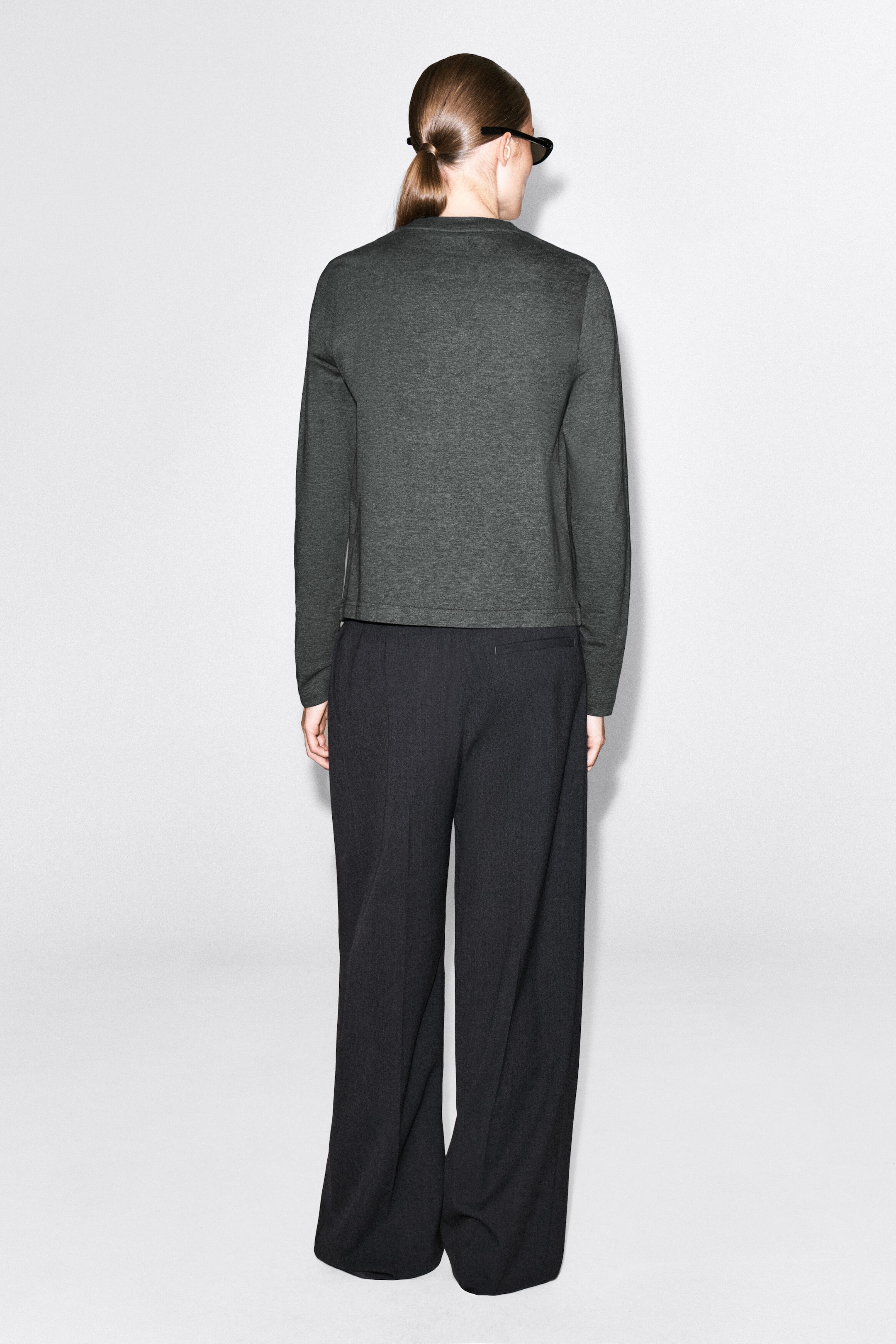 The model is wearing a clean-cut, long-sleeve T-shirt in dark gray, shown from the back. The top features a crew neck and a straight hem and is styled with dark wide-leg trousers.
