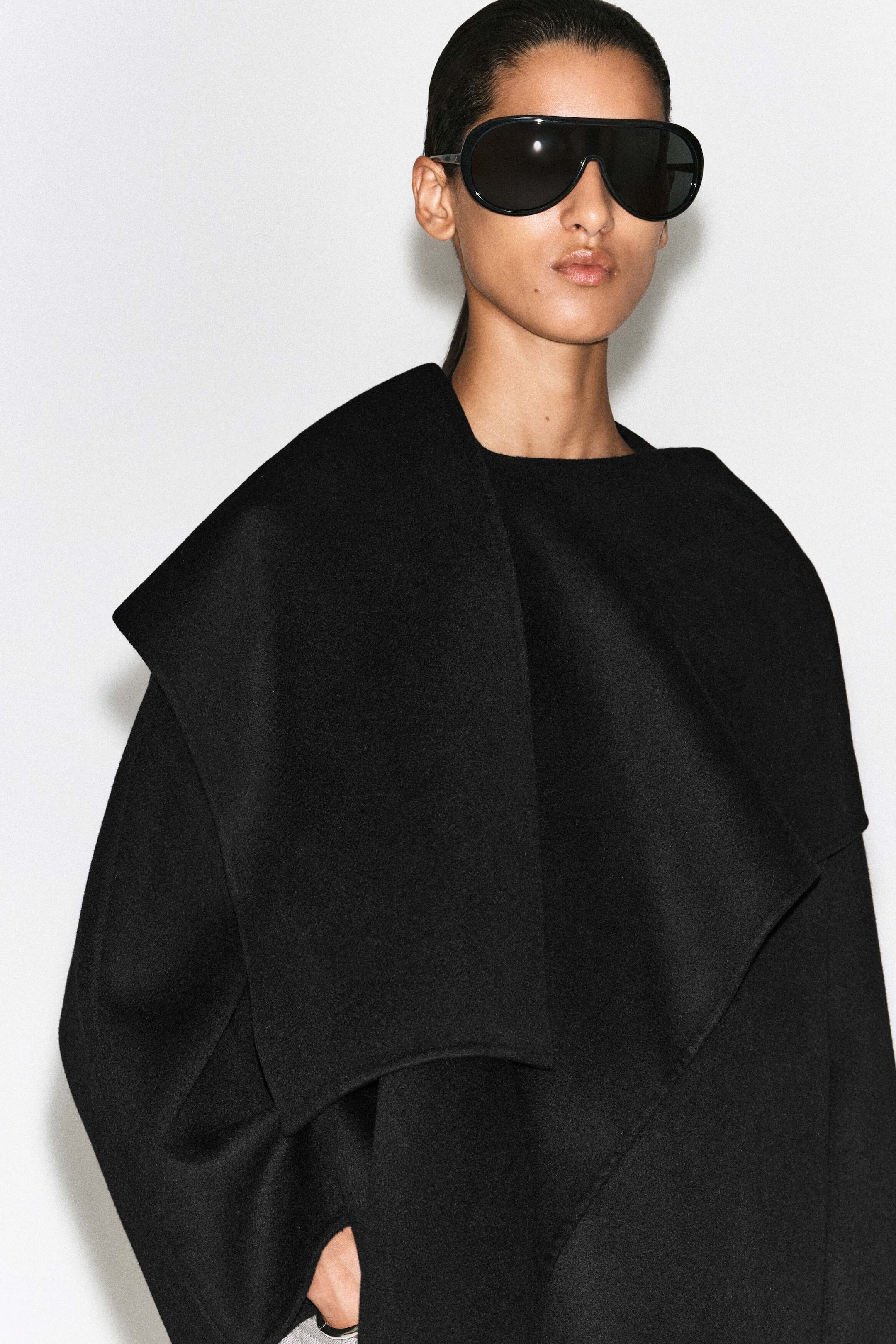 The double-faced wool-blend scarf jacket, shown in a close-up front view, features a draped, asymmetrical scarf-like collar that wraps over the shoulder and chest. The black jacket has a structured, oversized fit with clean-finished edges.
