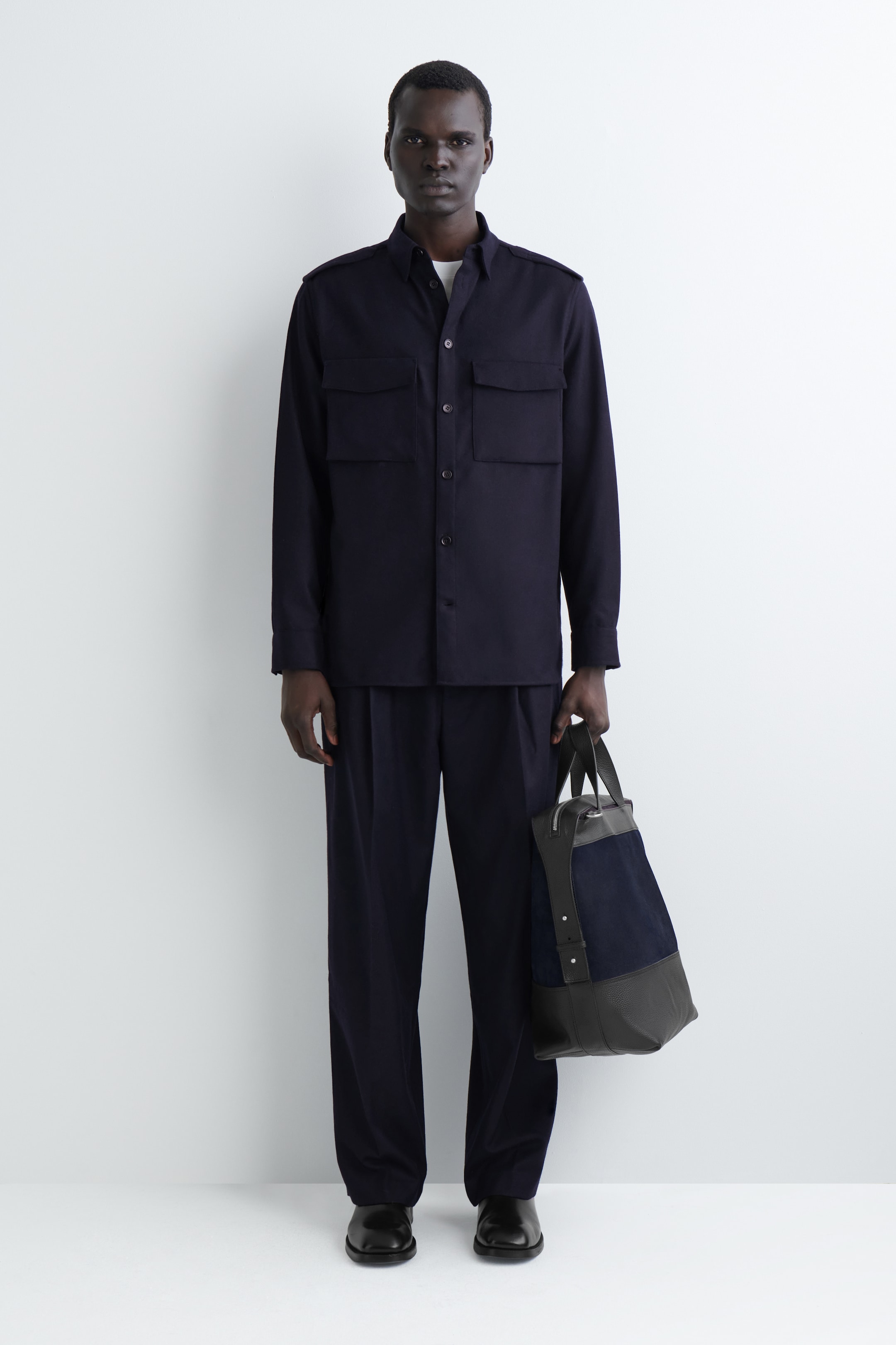 Ingrandisci l'immagine: Una camicia a maniche lunghe di colore blu navy scuro, con due tasche con patta sul petto e spalline sulle spalle, viene indossata con pantaloni a pieghe a gamba larga abbinati e scarpe eleganti nere. Una borsa tote in pelle grigio scuro a due toni e camoscio blu navy viene tenuta in mano, presentando l'outfit completo direttamente di fronte.