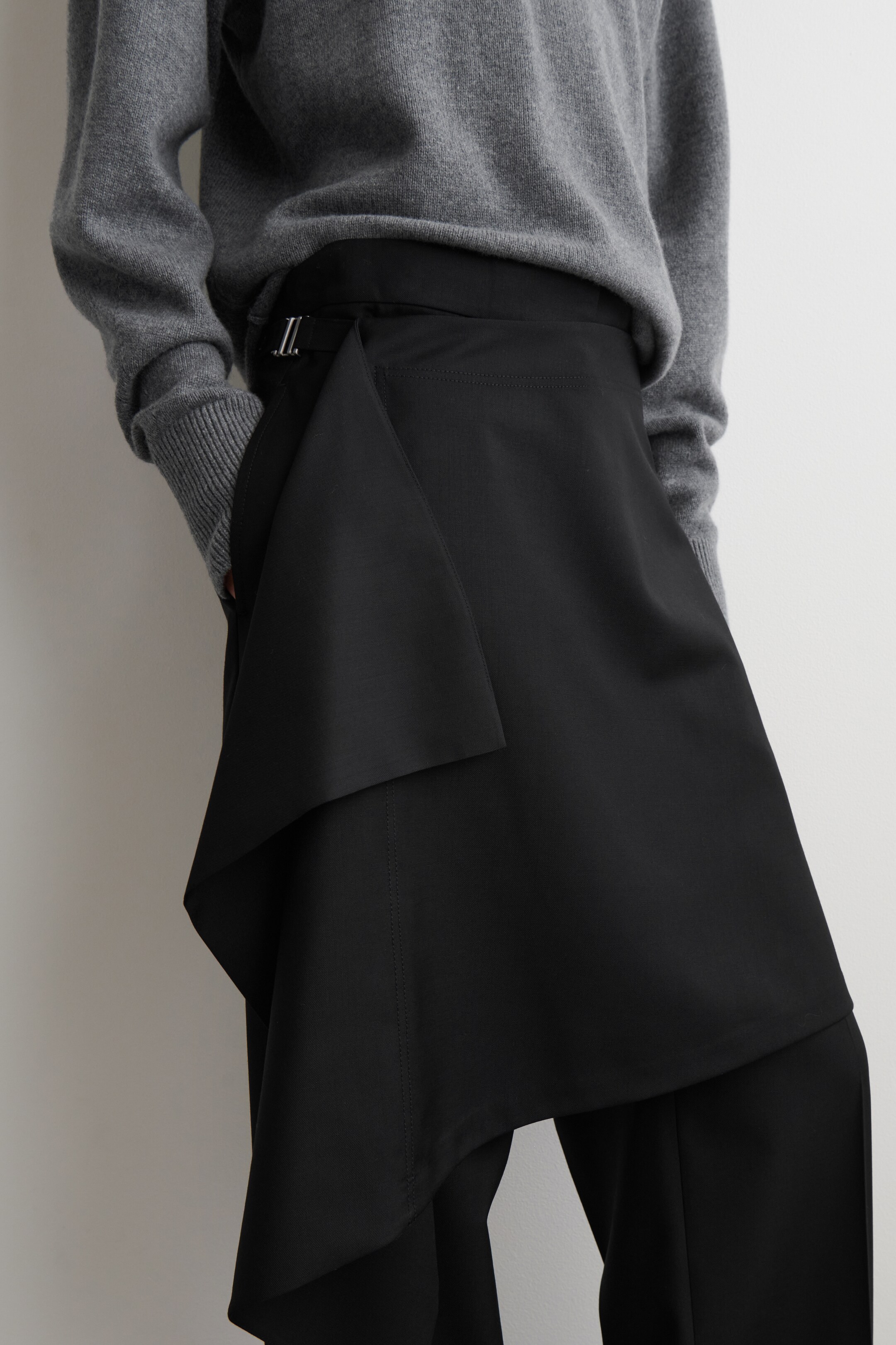 Agrandir l'image: Une jupe portefeuille asymétrique noire est portée par-dessus un pantalon noir, inclinée vers la gauche du porteur, affichant des panneaux de tissu superposés et une boucle argentée à la taille. Un pull en tricot gris est porté au-dessus de la jupe.