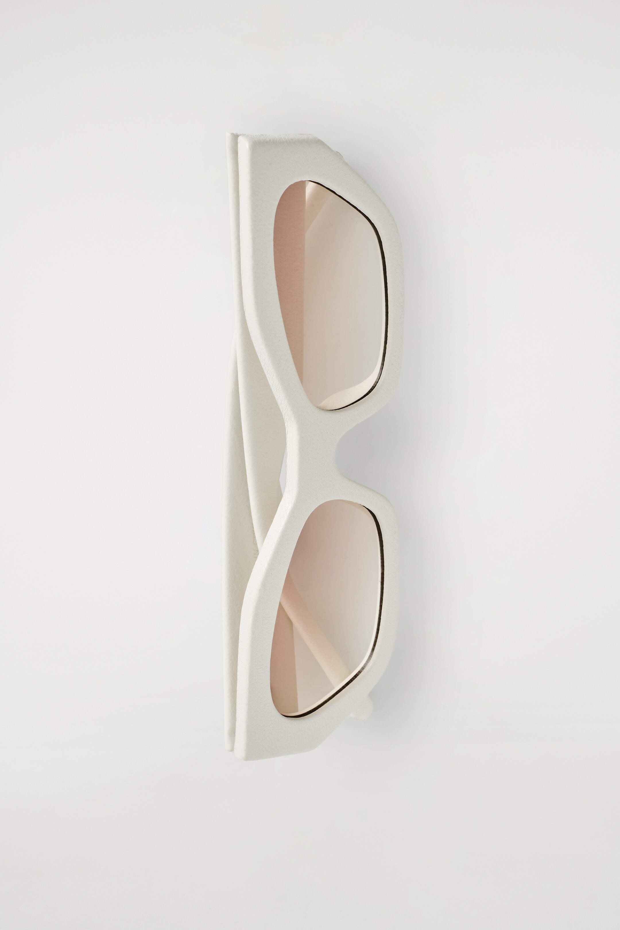 View larger image: A pair of sunglasses is lying on its side, featuring thick, off-white frames with a textured, matte finish. The rectangular lenses are tinted light pink, and the temples are folded inwards.
