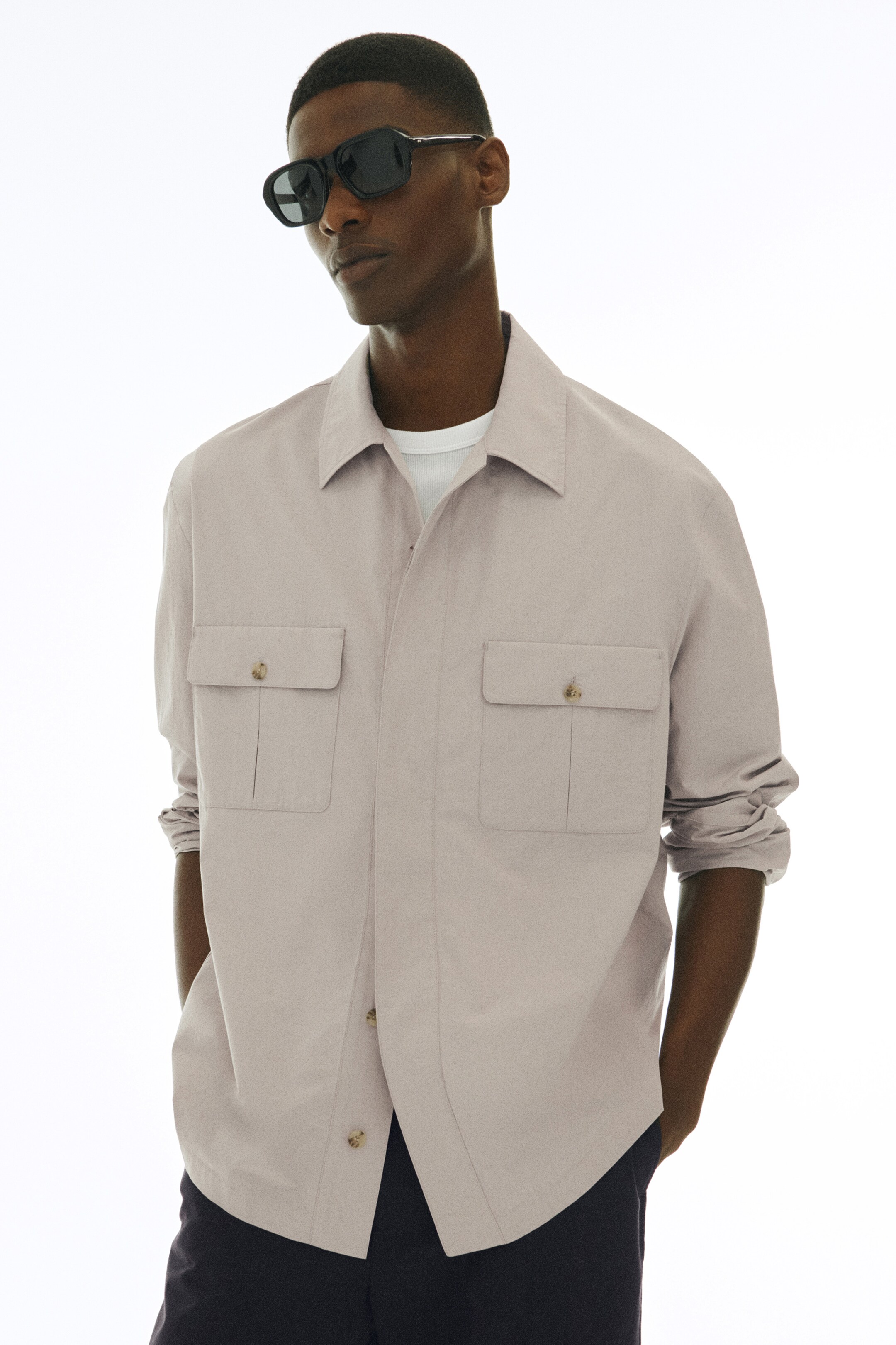 Größeres Bild anzeigen: Ein blassgraues Overshirt wird von vorne präsentiert, mit einer entspannten Passform, einem Umlegekragen und einer verdeckten Knopfleiste. Es ist mit zwei Brusttaschen mit Knopfklappen gestaltet und wird offen über einem weißen gerippten Unterhemd getragen, wobei die langen Ärmel an den Manschetten hochgekrempelt sind.