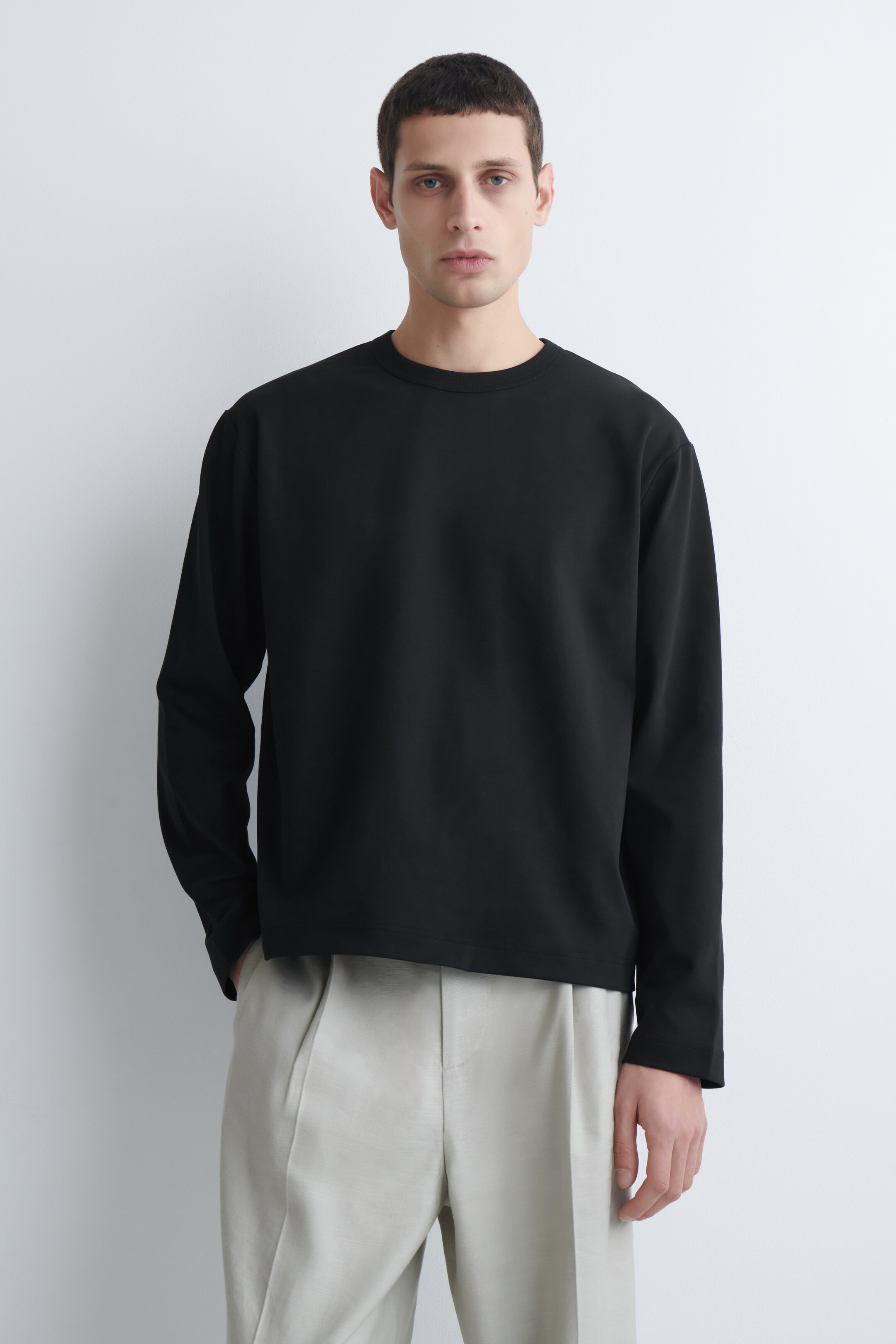 View larger image: A black long-sleeved crewneck top is being worn, facing forward. It features a relaxed fit with dropped shoulders and a straight hem with subtle side vents.