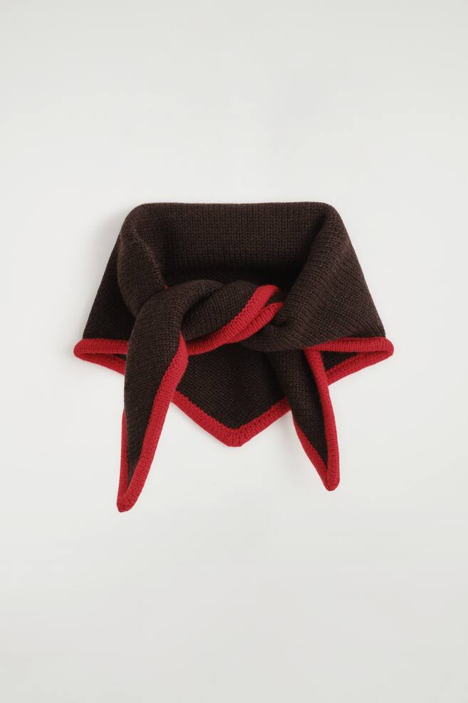 De driehoekige sjaal van merinowol met contrasterende afwerking toont een donkerbruine gebreide basis met een levendige rode contrastboord langs de randen. Het model wordt van voren weergegeven, gestyled met een losse knoop in het midden, waarbij de driehoekige punt naar beneden wijst en de twee uiteinden symmetrisch naar beneden vallen. - 1