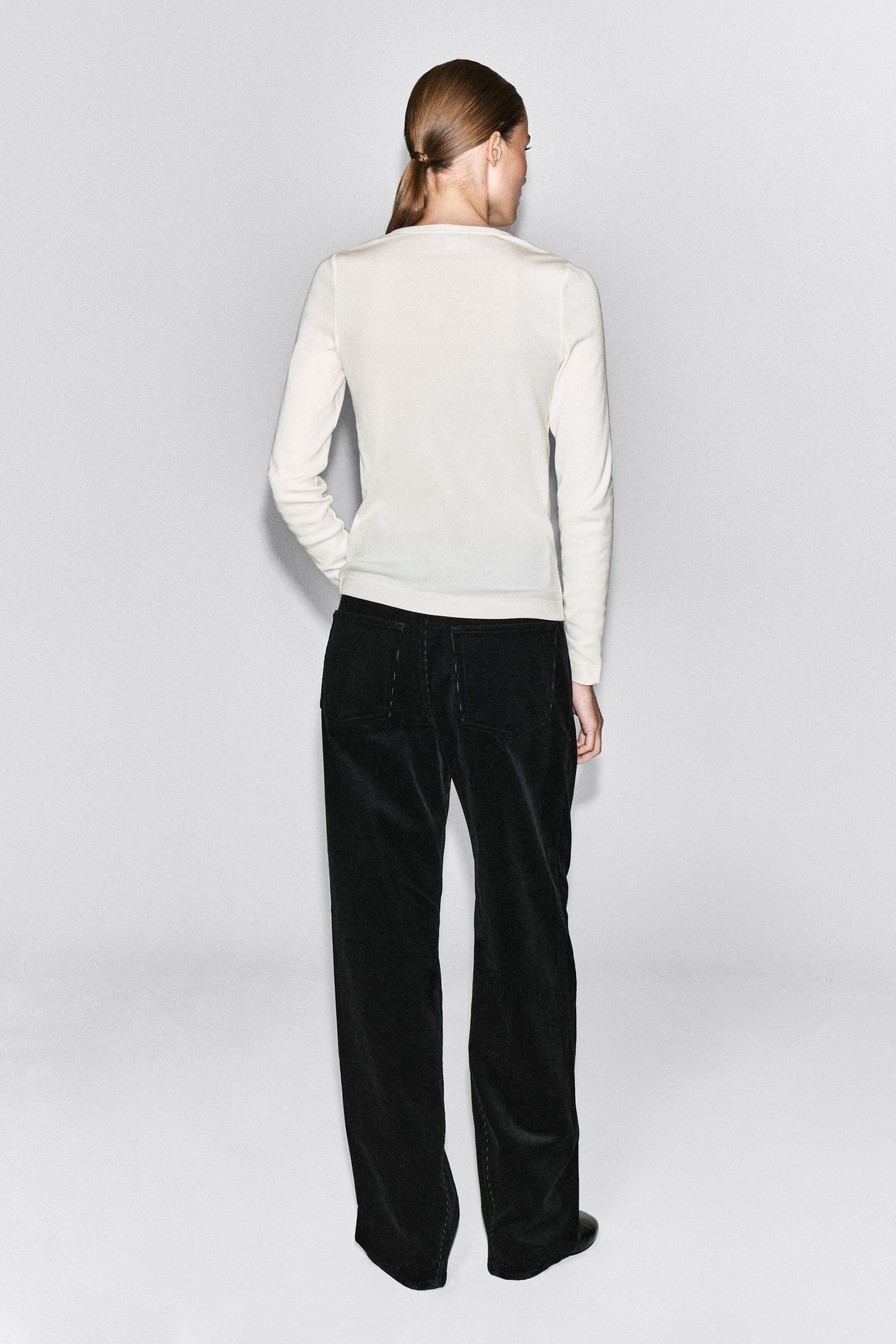A ribbed cotton henley top in off-white is being worn by the model, shown from the back. The long-sleeved top features a fine ribbed texture and is styled with black wide-leg trousers. A ribbed cotton henley top in off-white is being worn by the model, shown from the back. The long-sleeved top features a fine ribbed texture and is styled with black wide-leg trousers.
