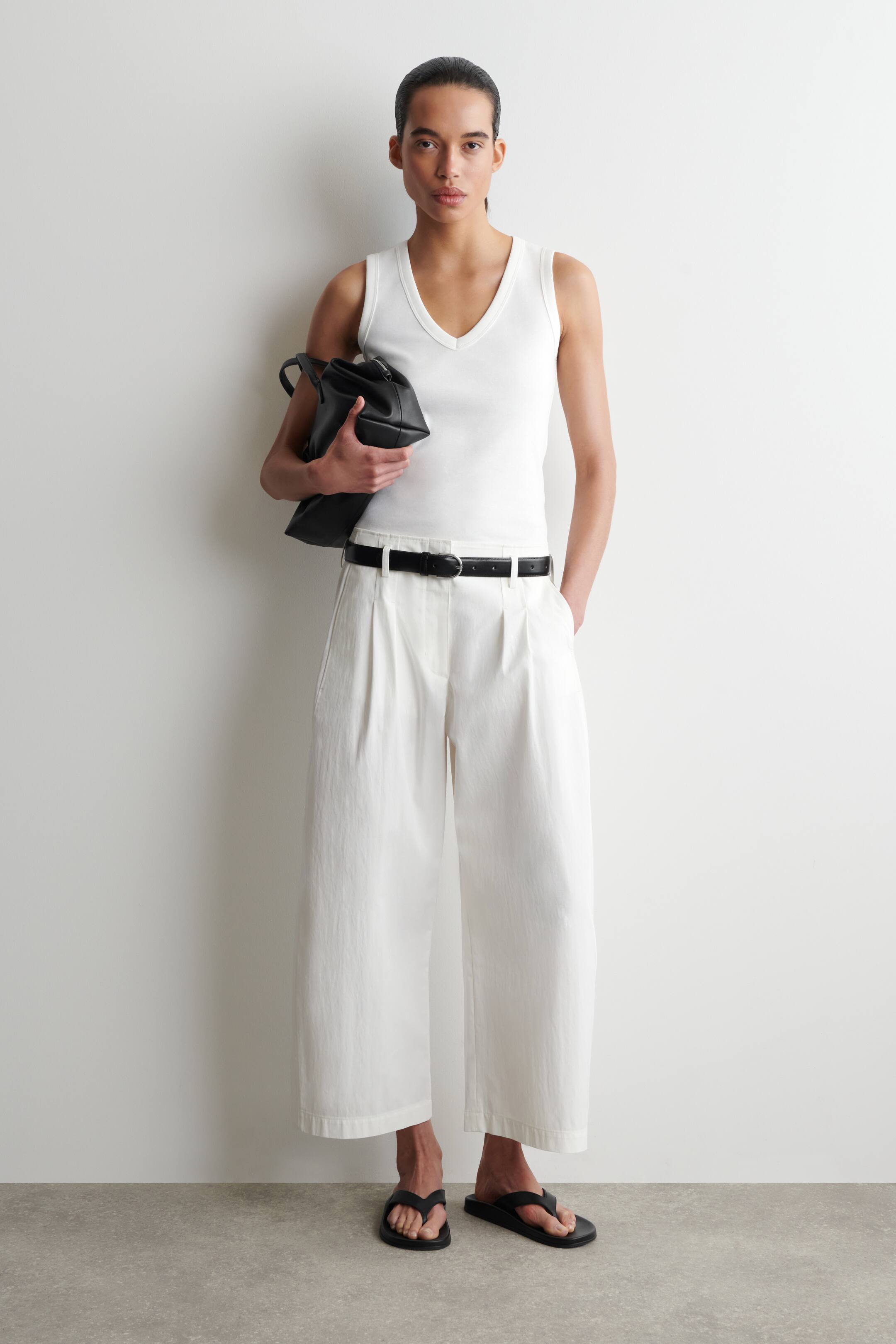 Ver imagen más grande: Se presenta una camiseta de tirantes blanca con cuello en V, ajustada al cuerpo. Se combina con pantalones blancos de pierna ancha con pliegues en la cintura y un cinturón negro, quedando los pantalones por encima del tobillo.