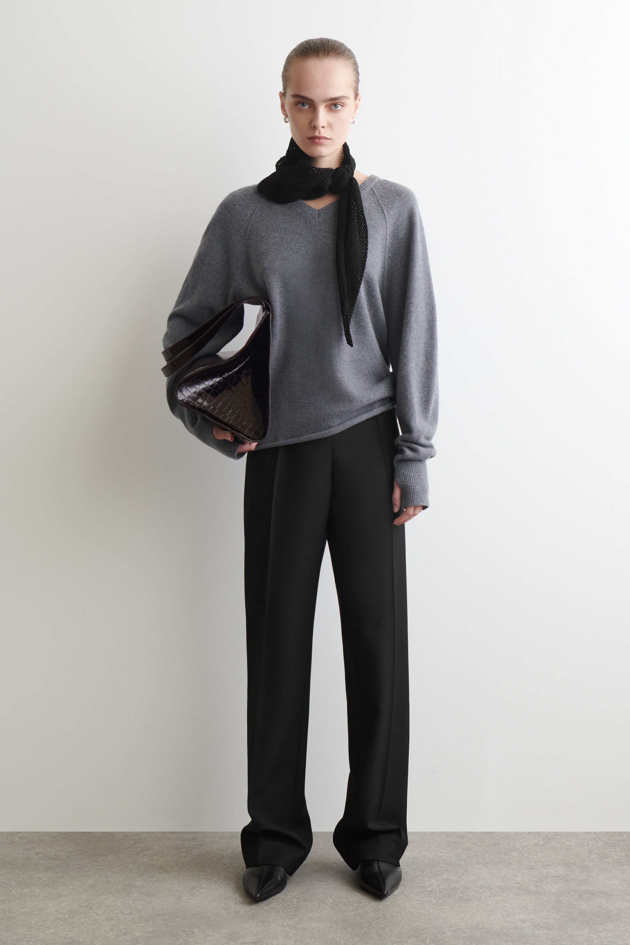 View larger image: A gray V-neck sweater with raglan sleeves and ribbed cuffs is being presented, paired with black wide-leg trousers and black pointed-toe shoes. A black triangular knit scarf is tied at the neck, and a dark brown crocodile-embossed handbag is being carried.