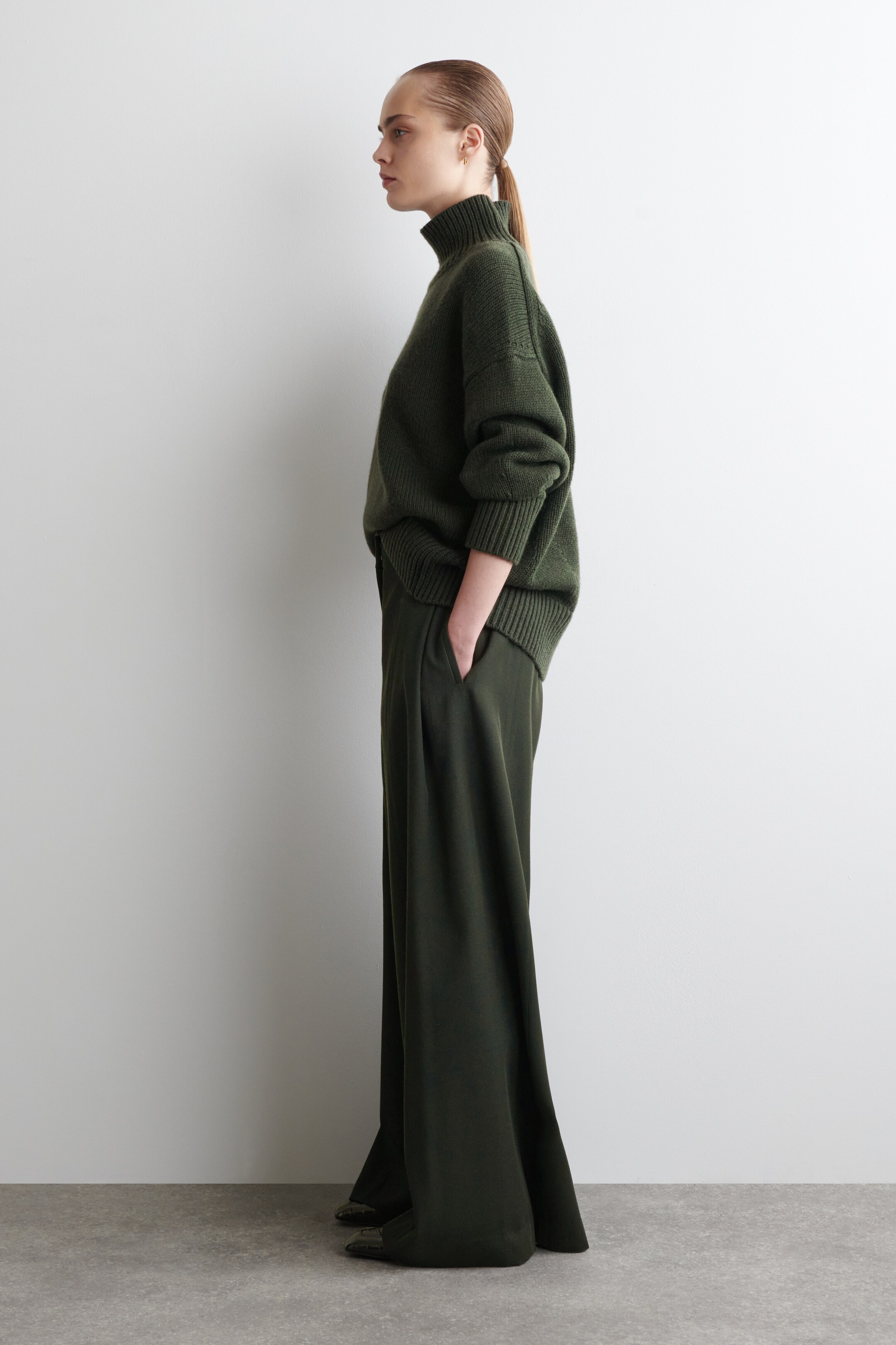 Visualizza immagine più grande: Un maglione a collo alto a maglia grossa di colore verde scuro con polsini a coste è indossato con pantaloni a gamba larga coordinati, visto di profilo laterale. Il maglione presenta una vestibilità rilassata con spalle scese, e i pantaloni mostrano pieghe e un drappeggio fluente.