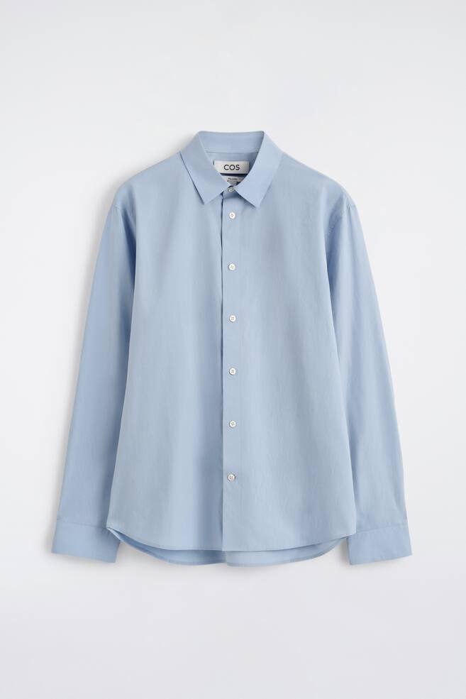A light blue long-sleeved shirt is displayed from the front, featuring a pointed collar, a full button-through placket with white buttons, long sleeves with buttoned cuffs, a relaxed fit, and a curved hem. - 1