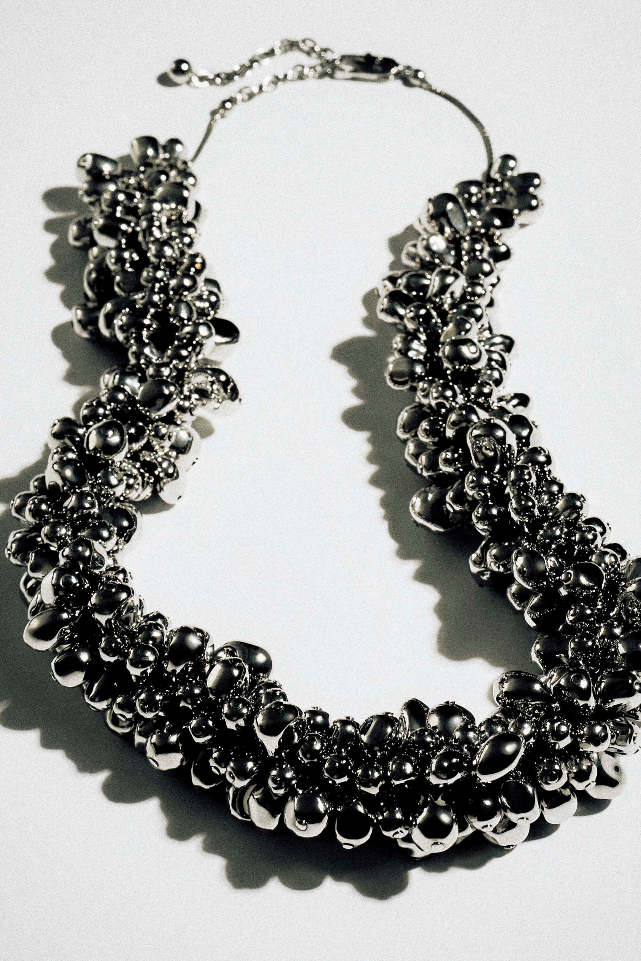 A silver-toned necklace features a dense, textured cluster of shiny, polished metallic spheres, shown lying flat in a front view. The necklace tapers at the top into a thin, delicate chain with a clasp closure. A silver-toned necklace features a dense, textured cluster of shiny, polished metallic spheres, shown lying flat in a front view. The necklace tapers at the top into a thin, delicate chain with a clasp closure.