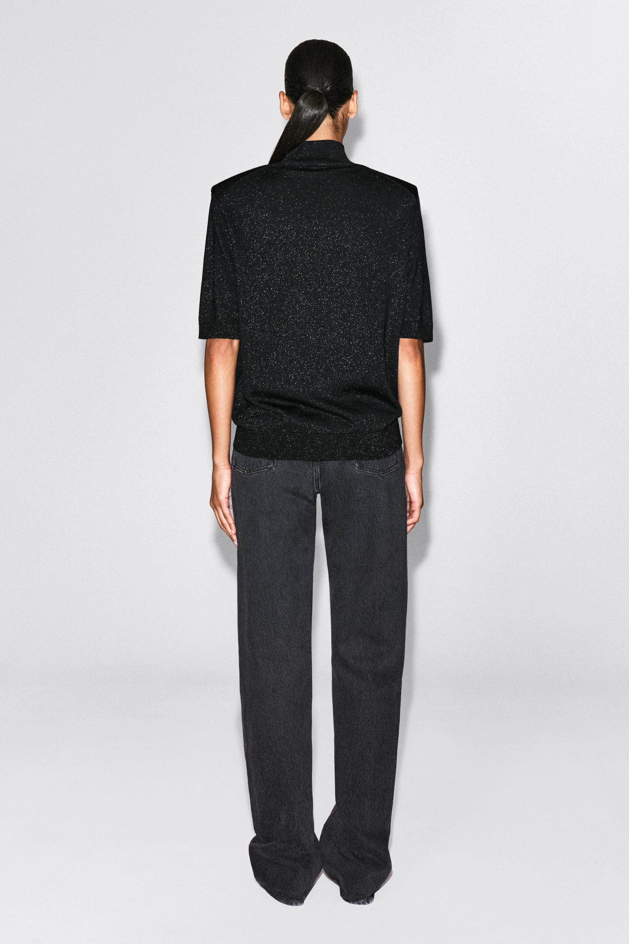 A black knit T-shirt featuring shimmering metallic flecks is shown from the back, highlighting its structured shoulders, high mock neck, and short sleeves. The model is wearing the top styled with dark grey straight-leg jeans. A black knit T-shirt featuring shimmering metallic flecks is shown from the back, highlighting its structured shoulders, high mock neck, and short sleeves. The model is wearing the top styled with dark grey straight-leg jeans.
