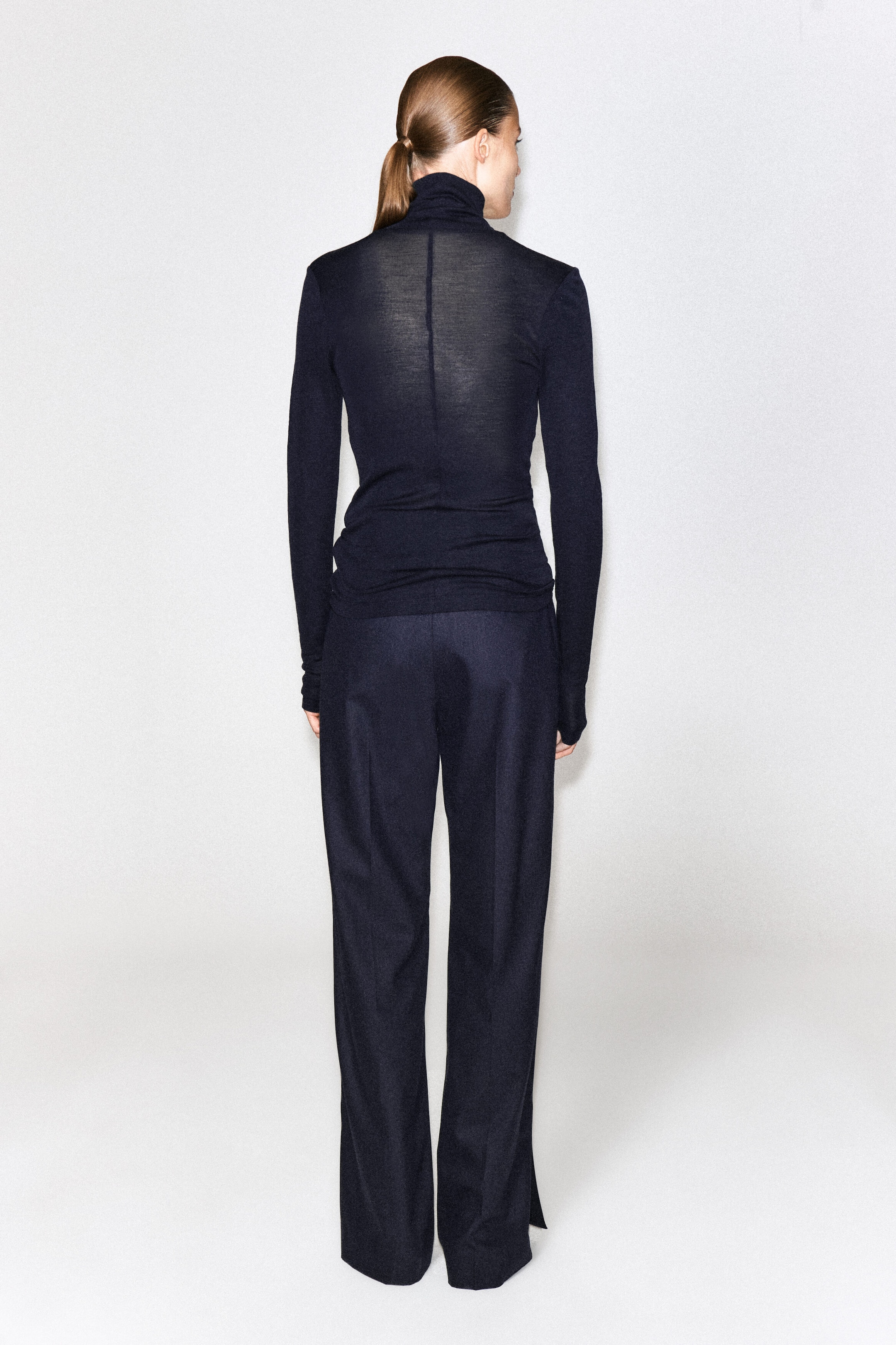 The model is wearing a dark navy, semi-sheer merino wool high-neck top with long sleeves and a center-back seam, styled with matching dark wide-leg trousers.
