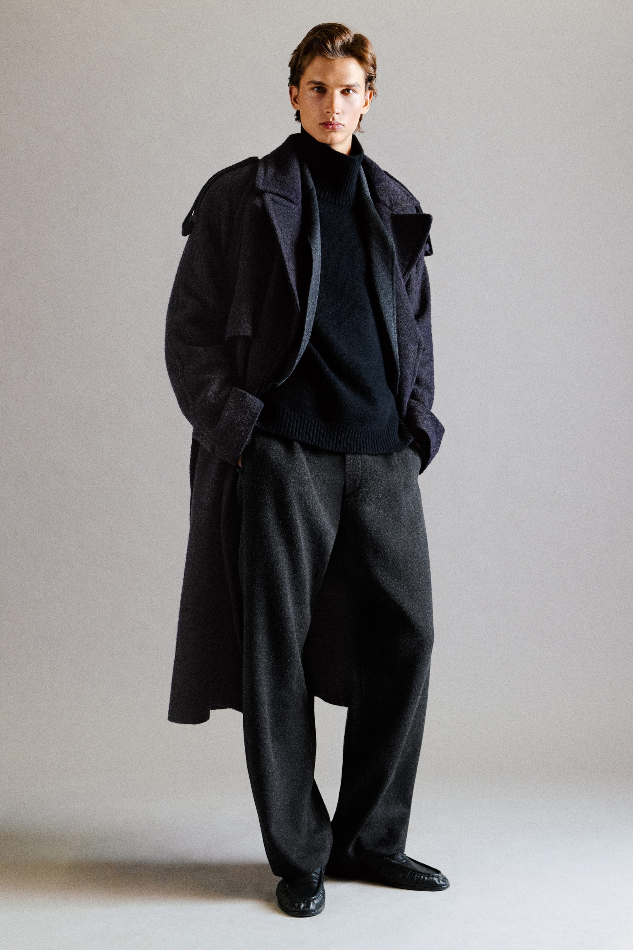 A model is wearing a relaxed-fit, dark charcoal wool-blend herringbone coat with epaulettes, layered open over a black turtleneck sweater in a front-facing view. The coat is paired with dark gray, wide-leg wool-blend trousers that slightly pool over black shoes. A model is wearing a relaxed-fit, dark charcoal wool-blend herringbone coat with epaulettes, layered open over a black turtleneck sweater in a front-facing view. The coat is paired with dark gray, wide-leg wool-blend trousers that slightly pool over black shoes.