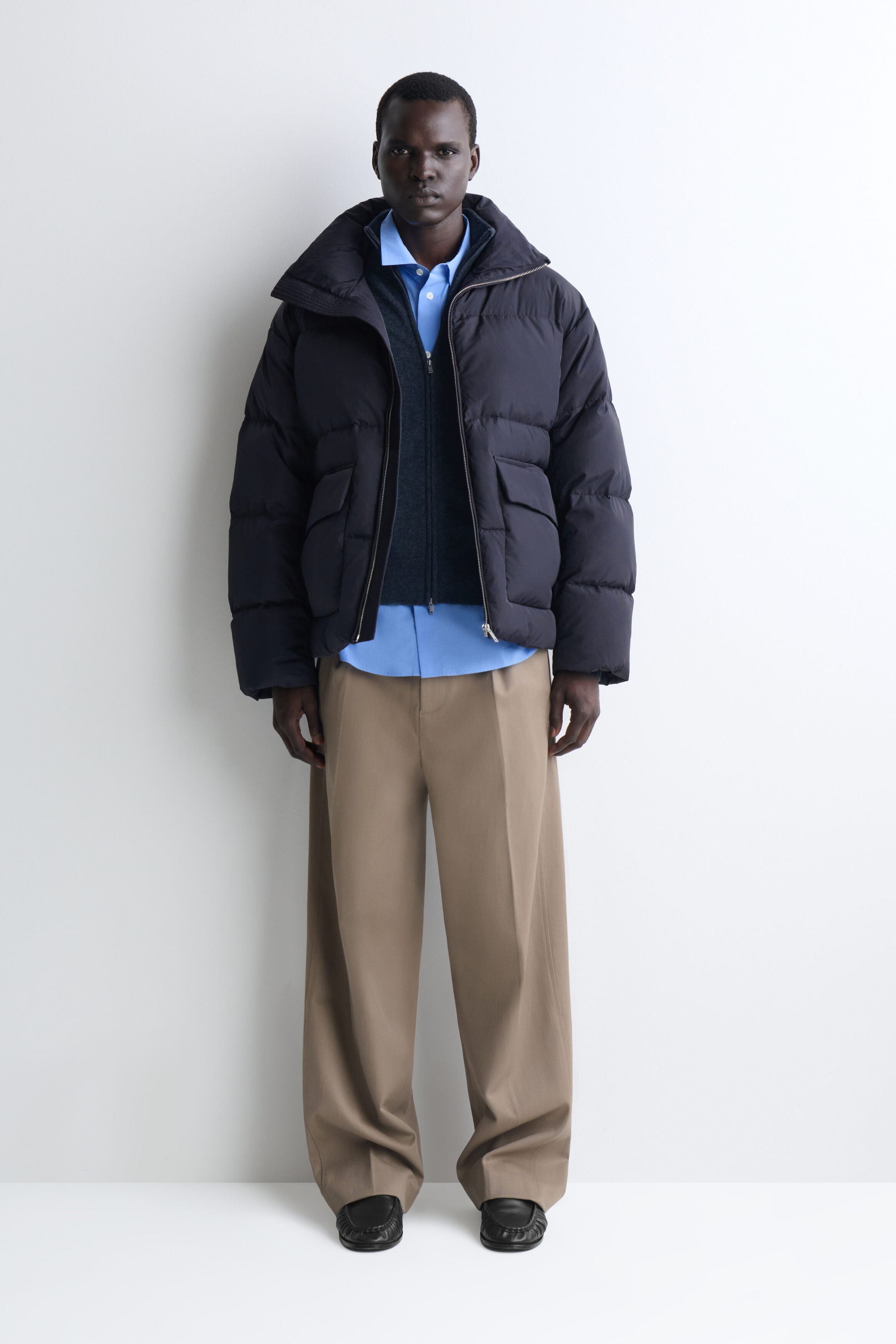 A dark navy funnel-neck nylon down puffer jacket is shown from the front, featuring horizontal quilting, large front flap pockets, and a wide, open collar. The jacket is layered over a dark gray zip-up cardigan and a light blue button-down shirt, showcasing its relaxed, structured fit.