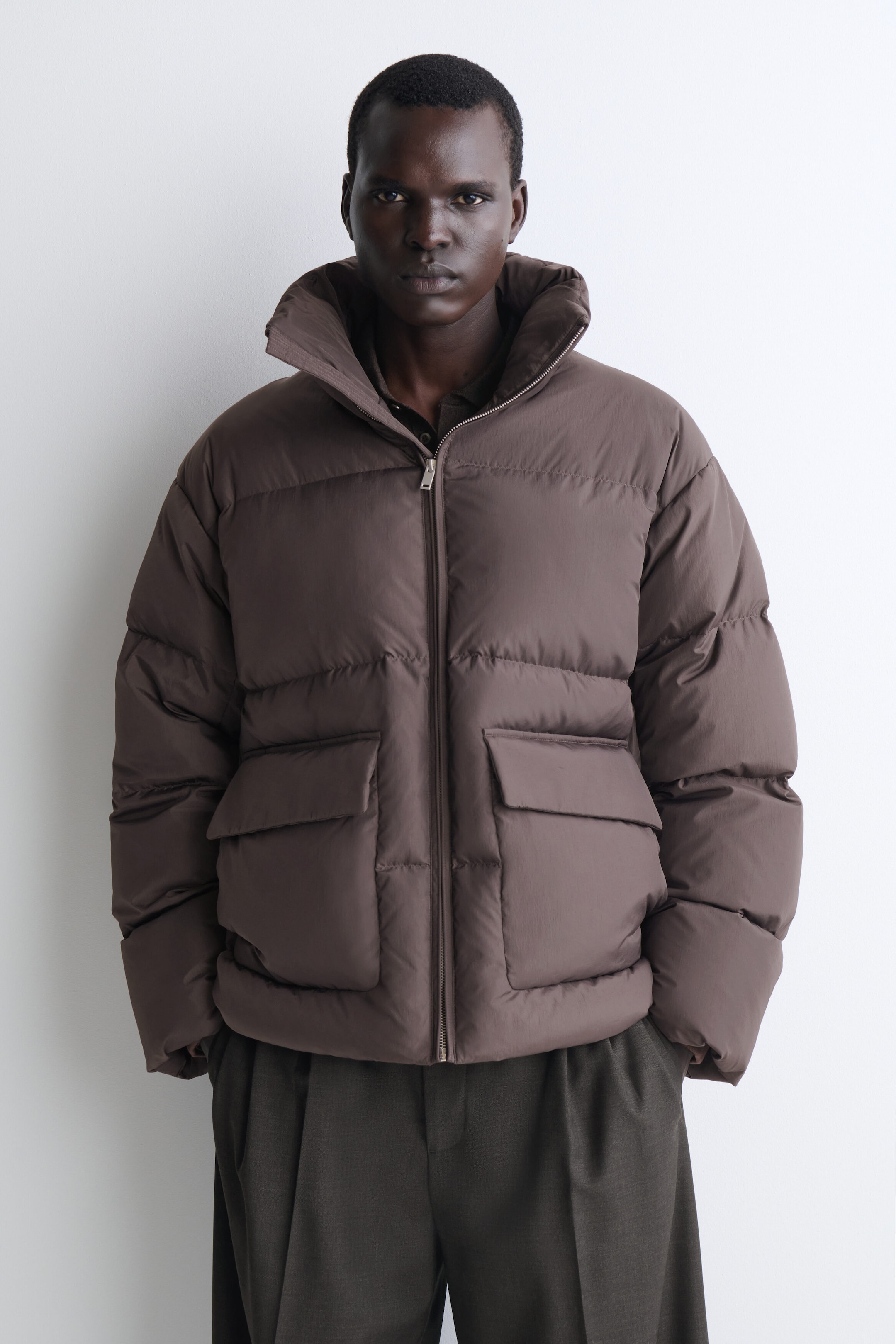 The brown funnel-neck nylon down puffer jacket is shown from the front, featuring horizontal quilted baffling, a high funnel neck, and a full-length front zipper. It showcases two large front pockets with flaps and a relaxed fit. The brown funnel-neck nylon down puffer jacket is shown from the front, featuring horizontal quilted baffling, a high funnel neck, and a full-length front zipper. It showcases two large front pockets with flaps and a relaxed fit.